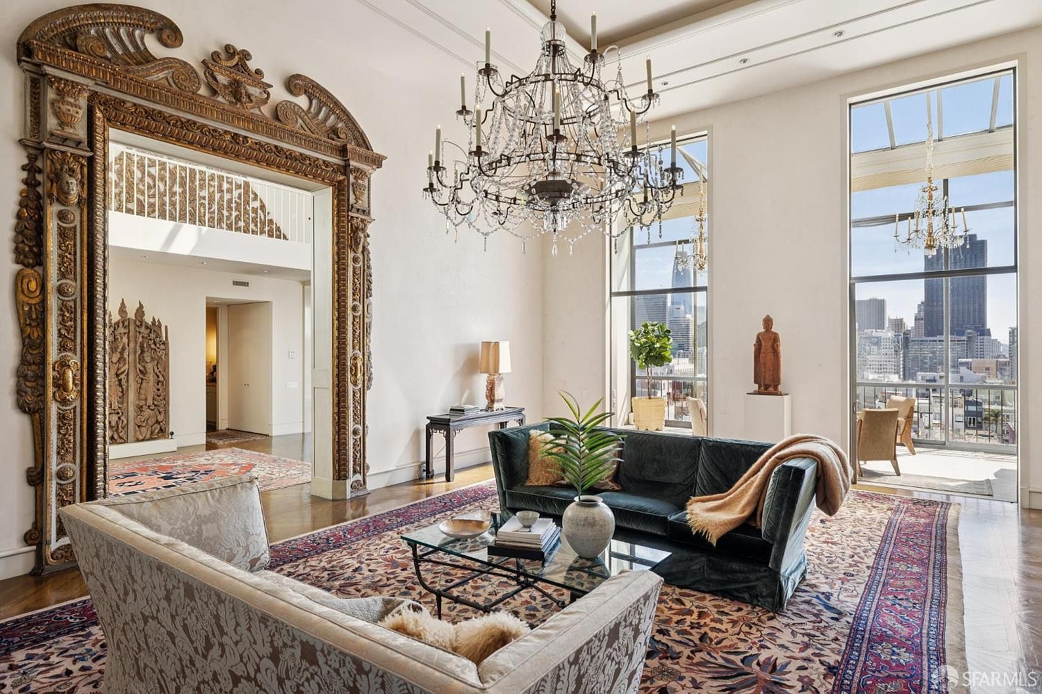 This is an interior shot of a luxurious living room featuring a large, ornate mirror frame, a crystal chandelier, and floor-to-ceiling windows offering a city view. The room is furnished with a dark green velvet sofa, a patterned armchair, and a glass coffee table, all set on an elaborate area rug. The overall impression is one of opulence and sophistication.