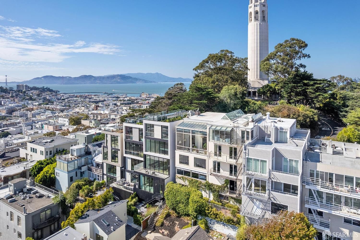 This aerial view showcases a modern multi-story residential building with a mix of glass and concrete facade, complemented by lush greenery and landscaping. The building features multiple balconies and rooftop terraces, offering stunning views of the city skyline and the bay. Coit Tower is visible in the background, adding to the property's unique appeal.