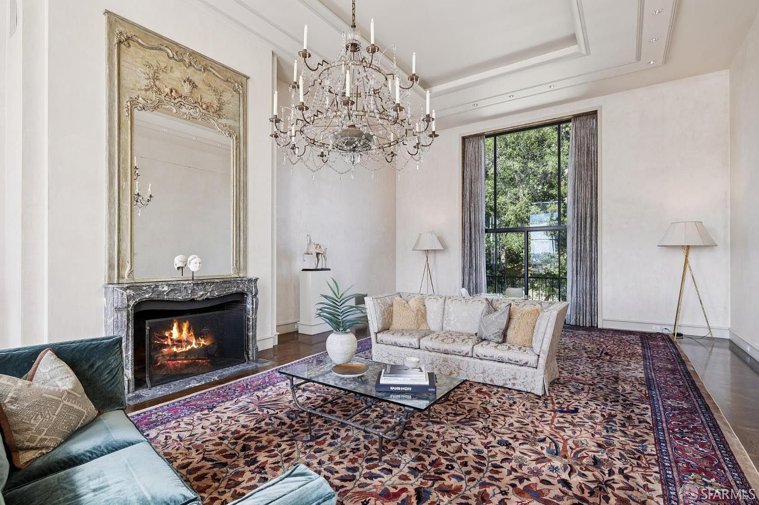 This is a luxurious living room featuring a grand chandelier, a fireplace with a decorative mirror above, and elegant furnishings. The room is bathed in natural light from a large window, and a patterned rug covers the hardwood floor. The overall impression is one of sophistication and classic elegance.