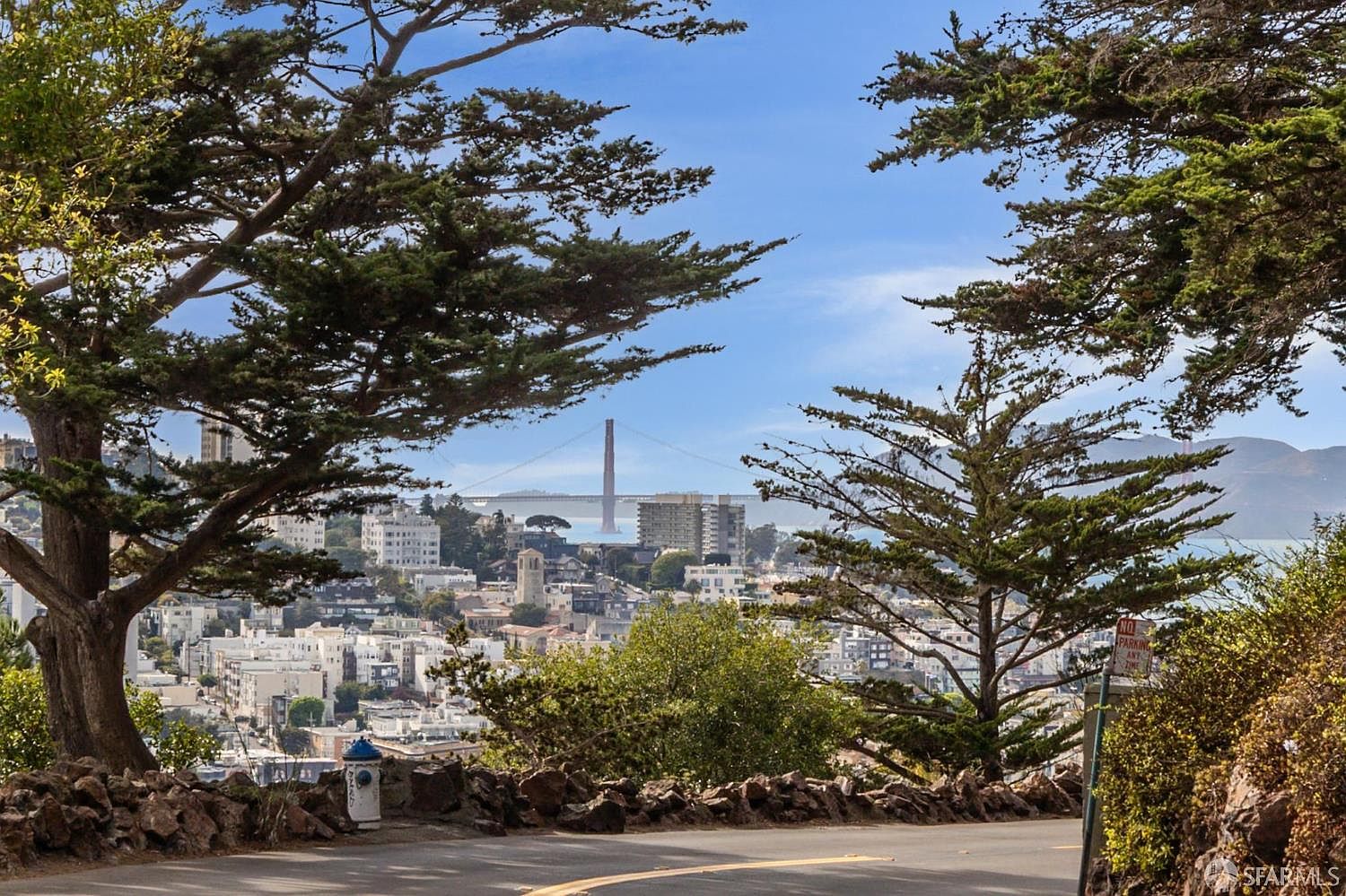This image showcases a scenic view of a city, possibly San Francisco, with the Golden Gate Bridge visible in the background. The foreground features lush trees framing the cityscape, adding depth and natural beauty to the scene. A winding road and stone wall suggest a vantage point offering panoramic views, enhancing the property's appeal.