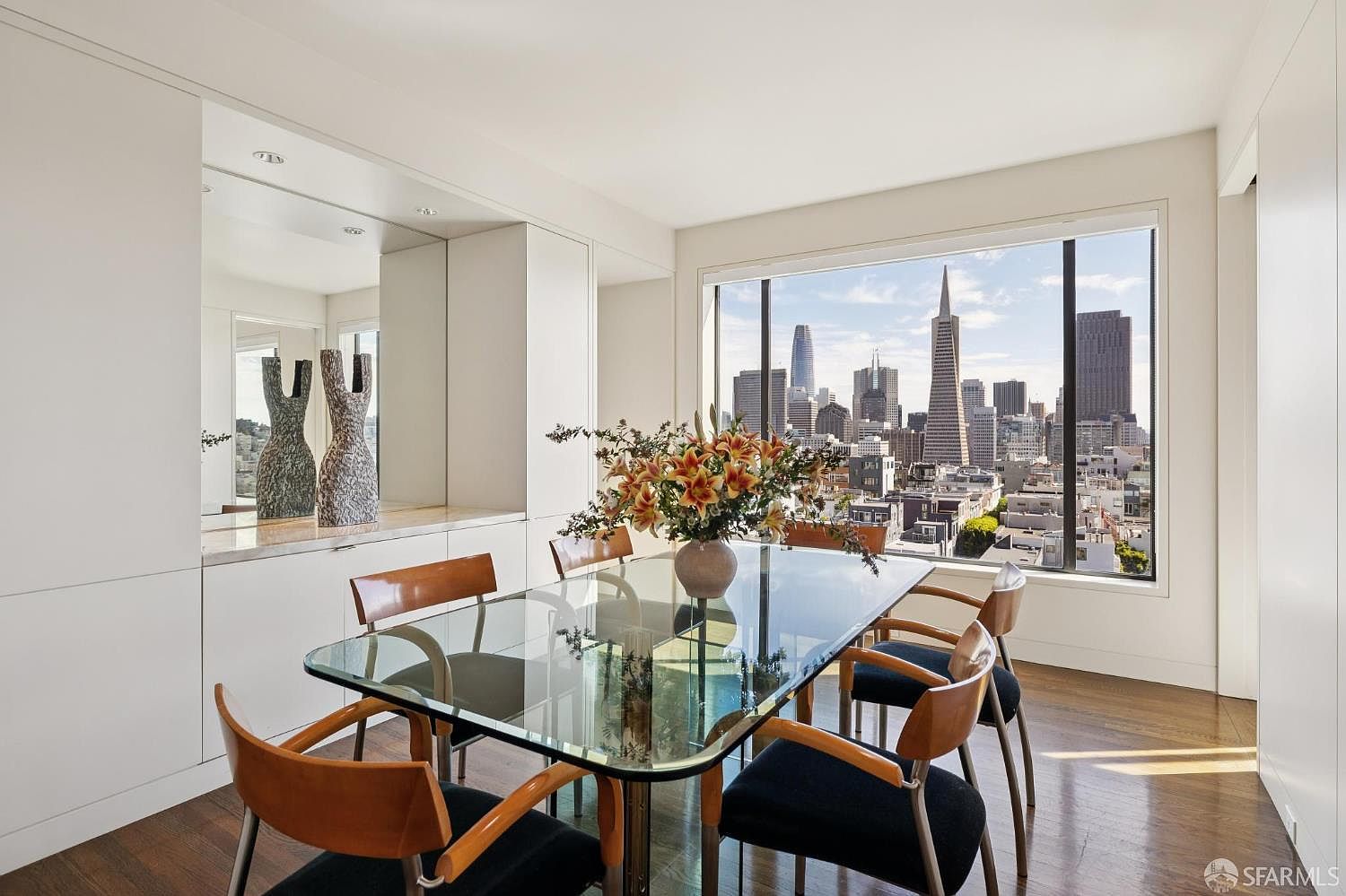 This dining room features a glass-top table surrounded by six chairs with wooden frames and dark cushions. A large window offers a stunning city view, including the Transamerica Pyramid. The room is bright and modern, with white walls and hardwood floors, creating an elegant and inviting space.