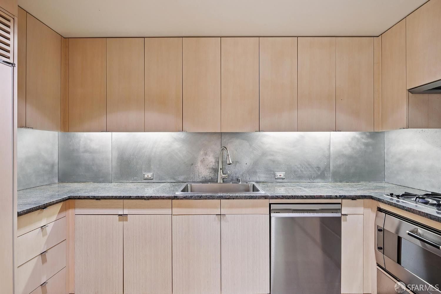 This is a well-lit kitchen featuring light wood cabinetry above and below a dark countertop. A stainless steel sink and faucet are centered beneath the upper cabinets, with a stainless steel backsplash. A stainless steel dishwasher is to the right of the sink, and a stainless steel oven is visible on the right edge of the frame.