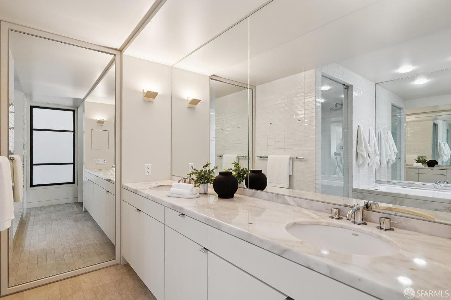 This is a well-lit primary bathroom featuring a long marble countertop with dual sinks and white cabinetry. A large mirror spans the length of the vanity, reflecting the space and enhancing the sense of openness. The bathroom also includes a separate shower area enclosed with glass, contributing to a clean and modern aesthetic.
