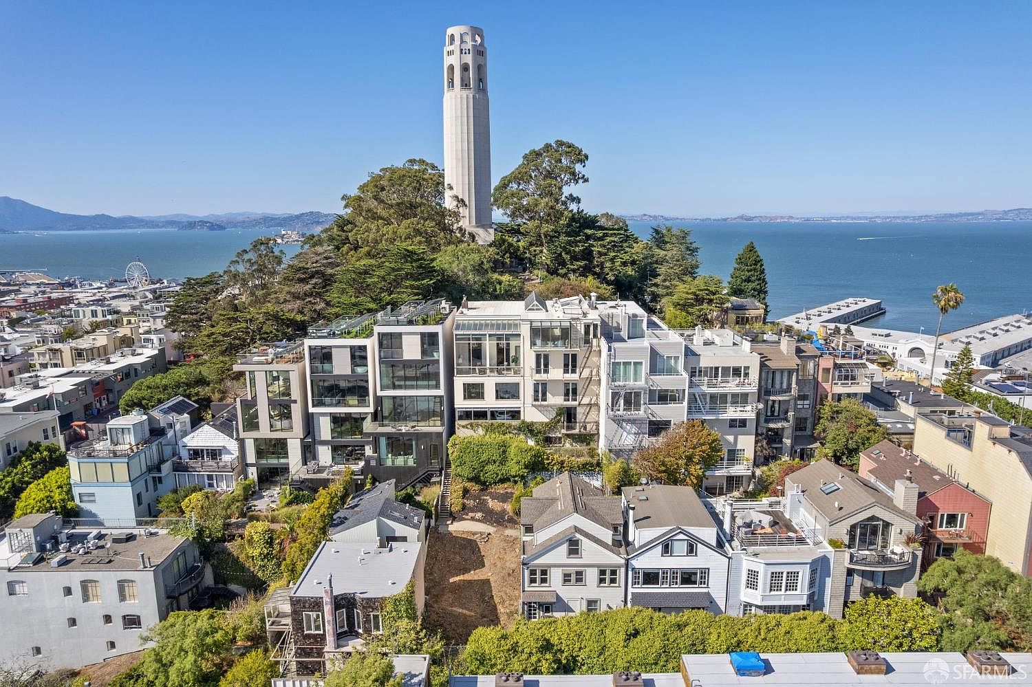 This aerial shot showcases a cluster of modern homes nestled on a hillside, with the iconic Coit Tower prominently featured in the background. The properties boast contemporary architectural designs, blending seamlessly with the lush greenery and offering stunning views of the bay. The overall impression is one of luxury and exclusivity in a prime location.