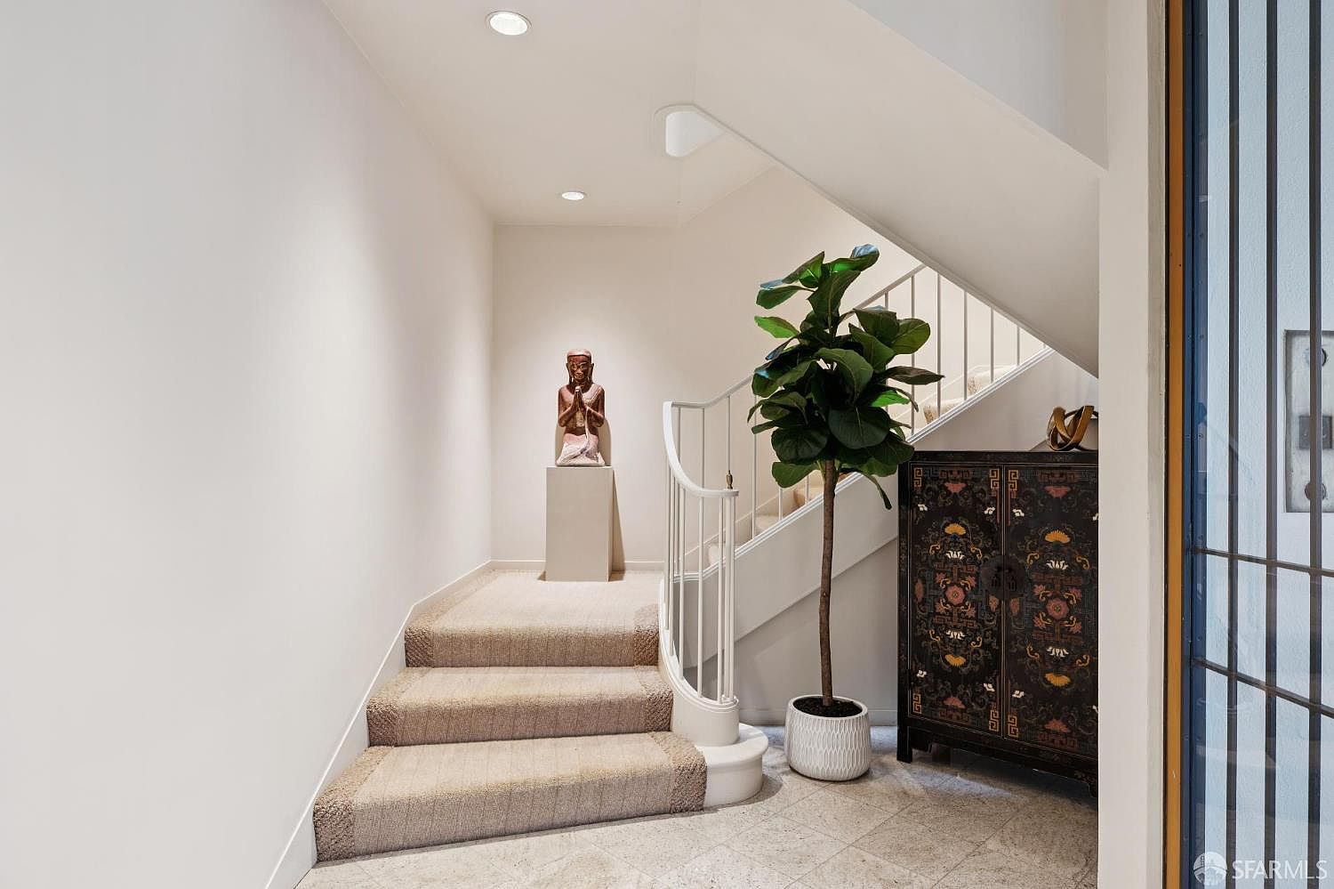 This interior shot showcases a well-lit hallway and staircase. The stairs are carpeted, leading up to a landing with a statue on a pedestal. A decorative cabinet and a potted plant add visual interest to the space, creating a welcoming and stylish transition area.