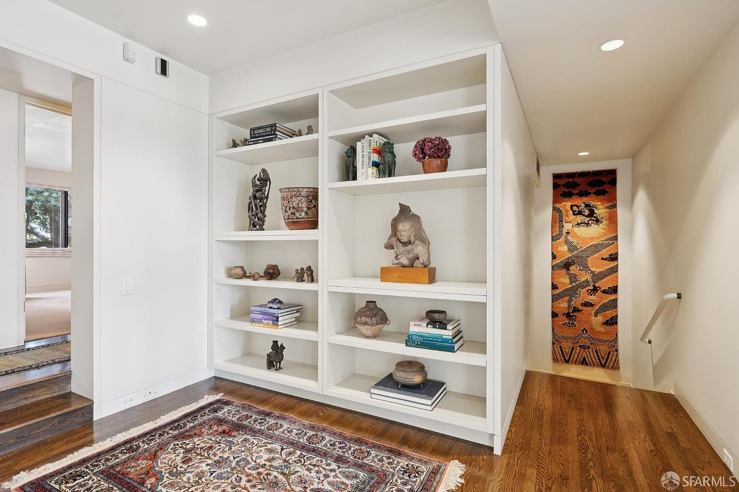 This interior shot showcases a hallway featuring built-in white bookshelves filled with decorative items and books. The hardwood flooring adds warmth, complemented by an ornate area rug. A glimpse into an adjacent room and a decorative wall hanging add depth and character to the space.