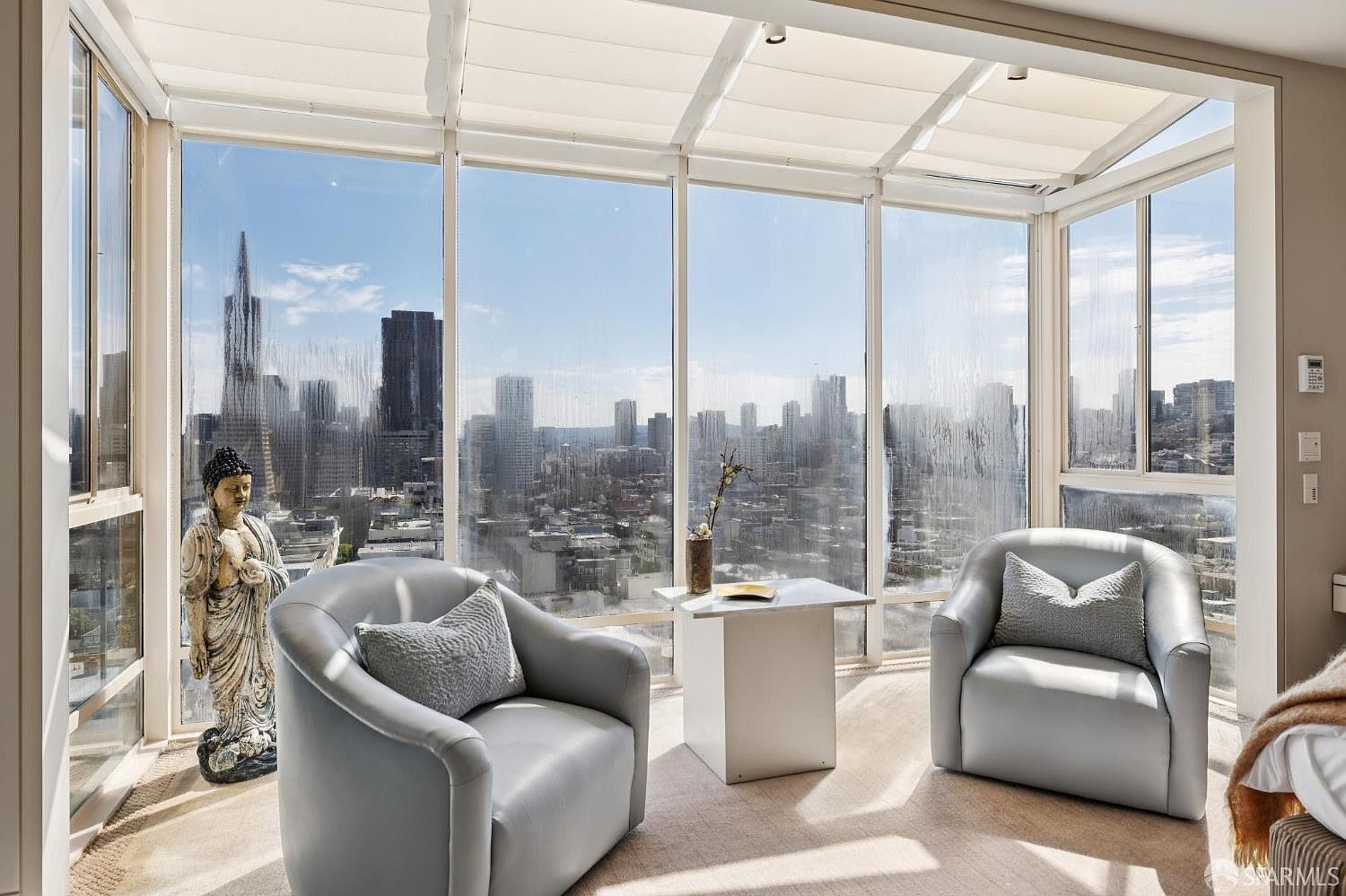 This sunroom features floor-to-ceiling windows offering panoramic city views. Two light gray armchairs with decorative pillows are arranged around a small white table, creating a cozy seating area. A Buddha statue adds a touch of serenity to the space, which is bathed in natural light.
