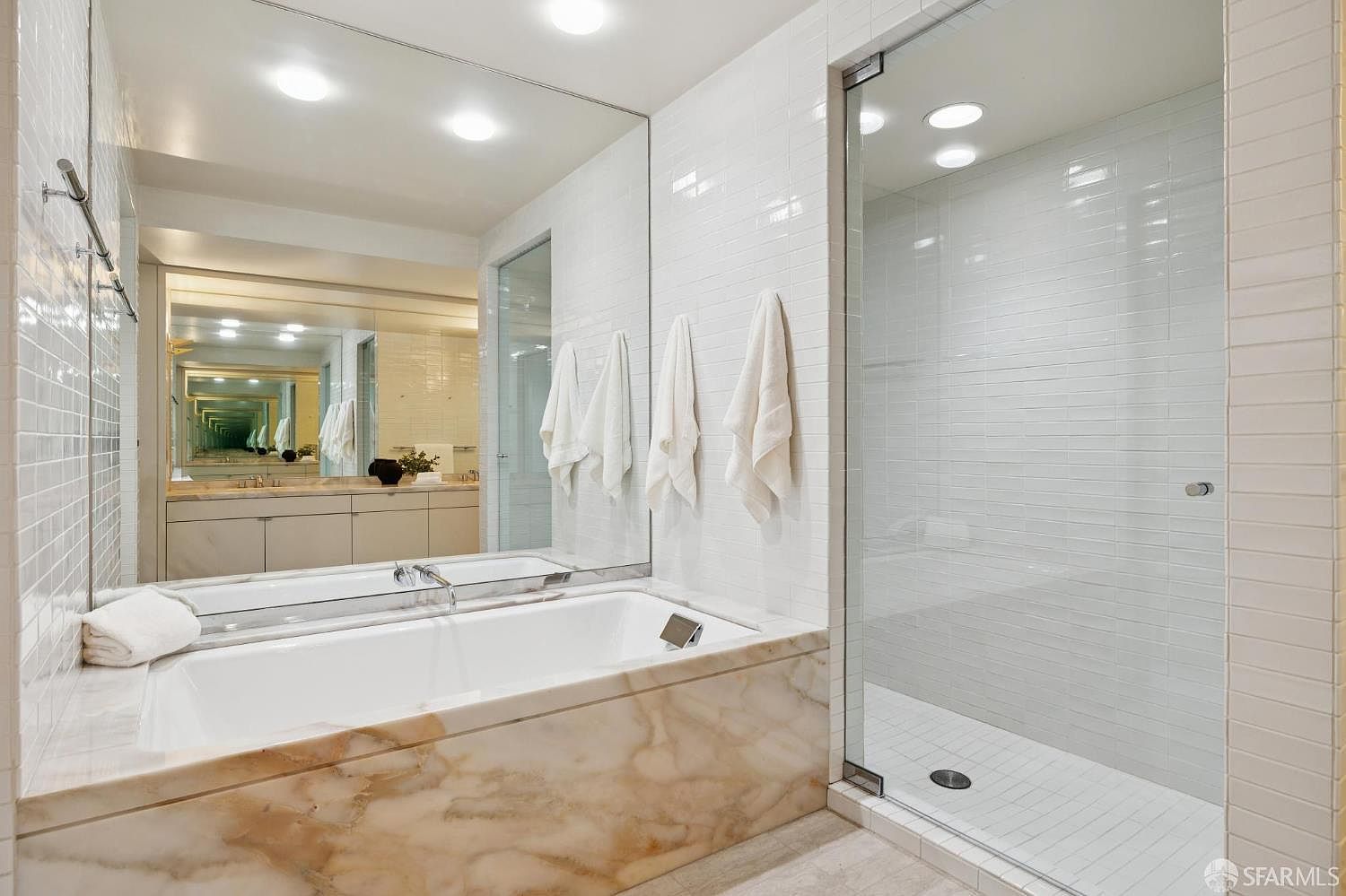 This is a luxurious primary bathroom featuring a marble-clad bathtub with a white interior, complemented by a glass-enclosed shower with white tiled walls. A large mirror reflects the vanity area, creating an illusion of spaciousness and enhancing the natural light. The overall impression is one of elegance and tranquility.