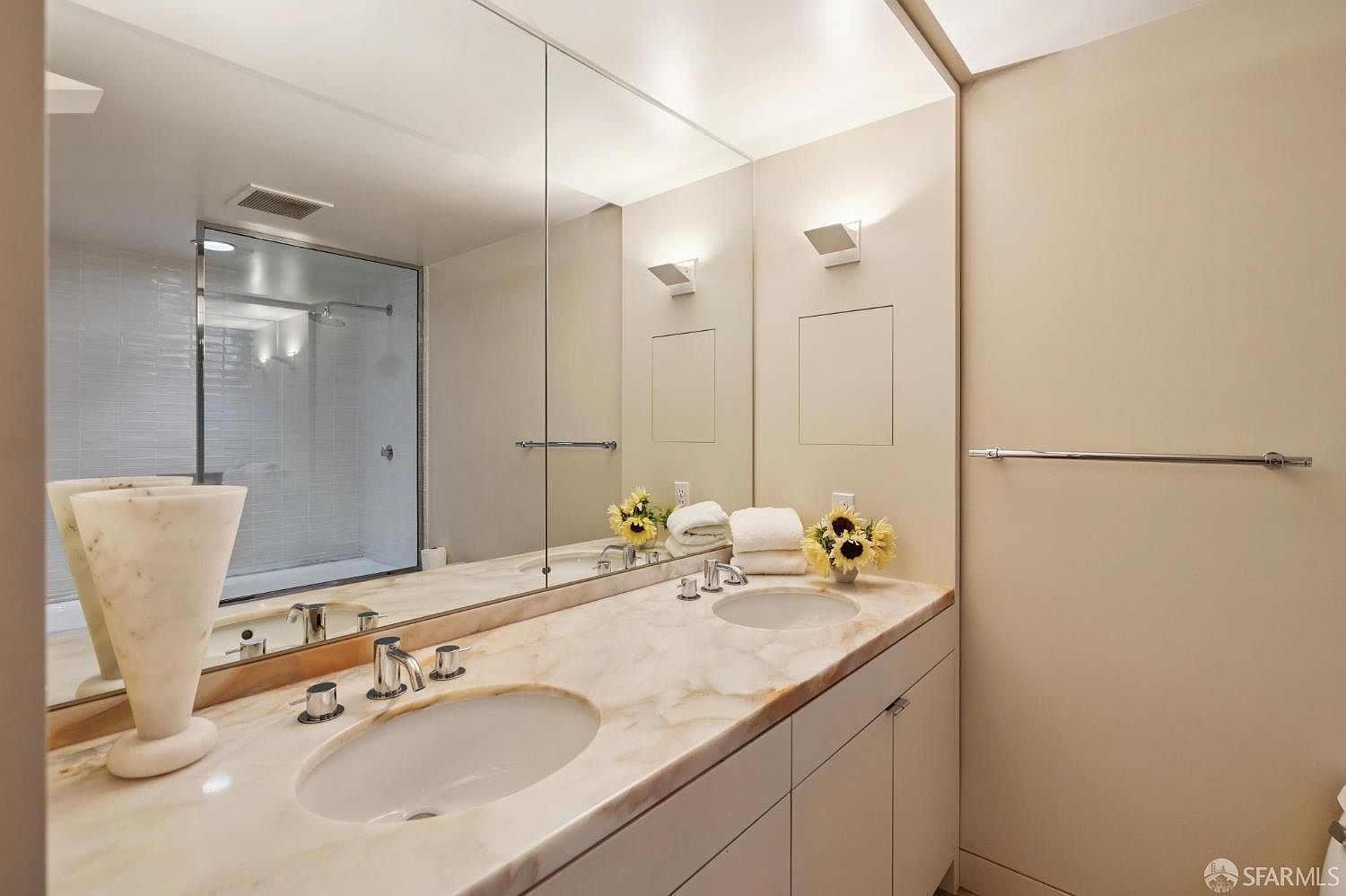 This is a well-lit primary bathroom featuring a double vanity with a marble countertop and dual sinks. A large mirror spans the length of the vanity, reflecting a glass-enclosed shower. The walls are painted in a neutral tone, and modern sconces provide additional lighting, creating a clean and luxurious atmosphere.
