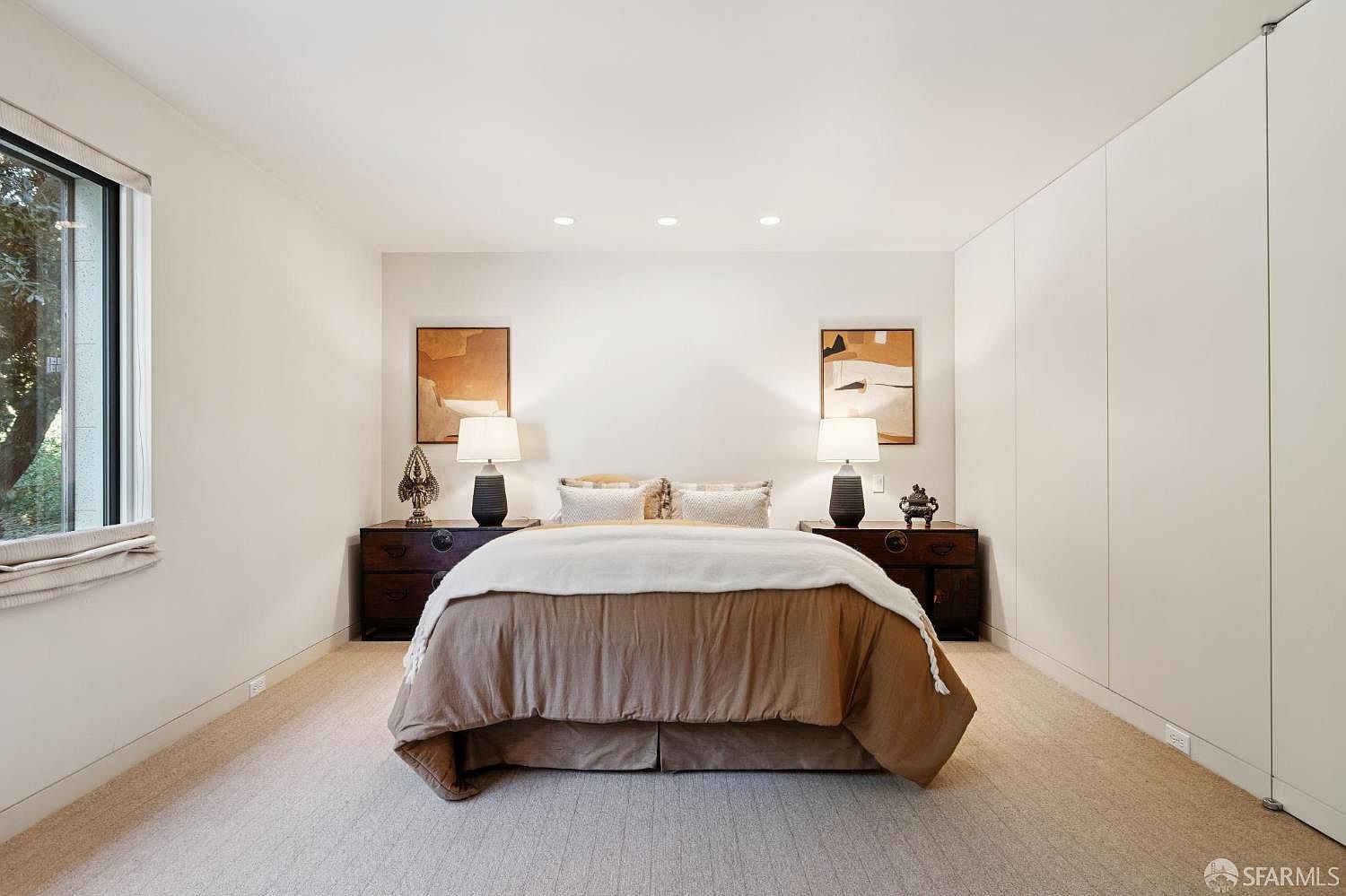 This is a well-lit primary bedroom featuring a neutral color palette. The room includes a large bed with a brown duvet and white throw, flanked by matching dark wood nightstands with lamps and decorative objects. Artwork hangs above each nightstand, and a large window provides natural light, while built-in closets offer ample storage.