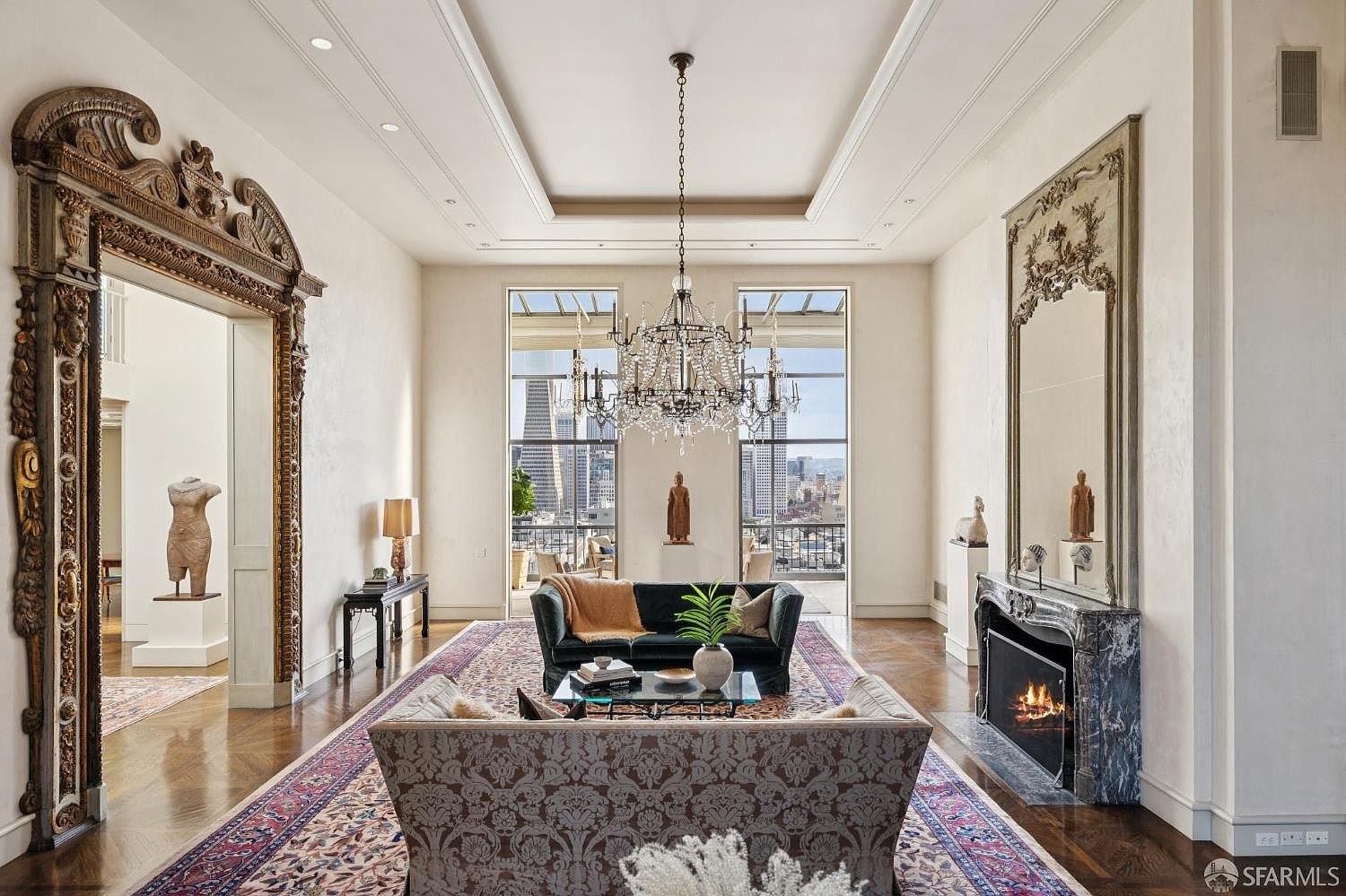 This is a luxurious living room featuring a large chandelier, floor-to-ceiling windows with city views, and ornate mirrors. The room is furnished with elegant sofas and a patterned rug, creating a sophisticated and inviting atmosphere. A fireplace adds warmth and character to the space.
