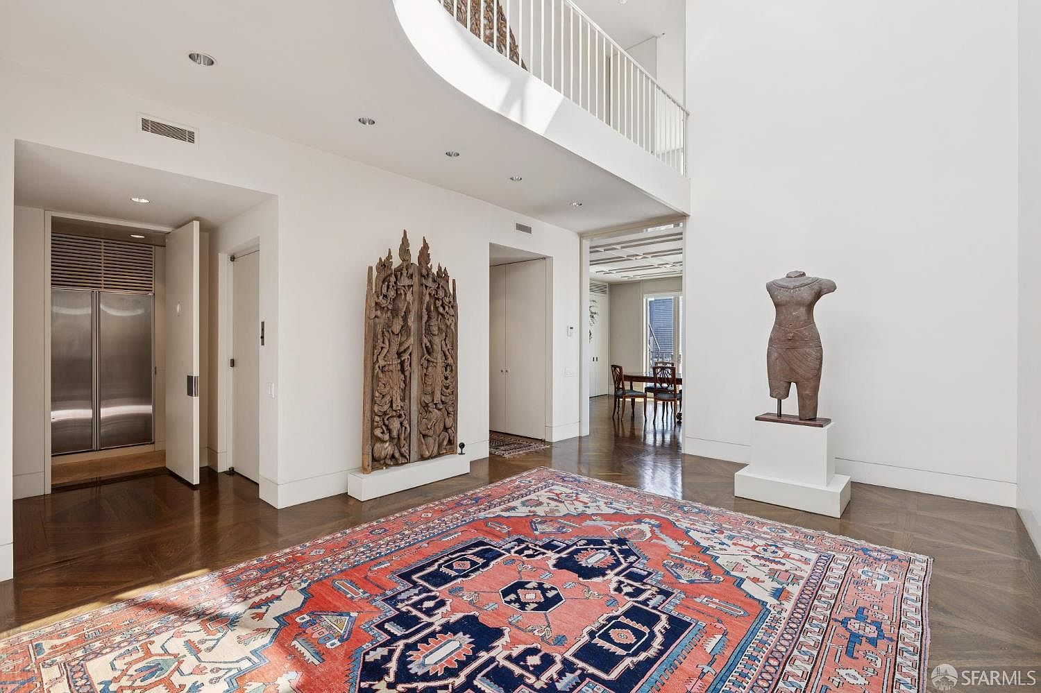 This is an interior shot of a grand hallway featuring dark hardwood floors and white walls. A large, colorful rug anchors the space, while a wooden sculpture and a torso statue add artistic elements. A staircase with white railings is visible in the background, contributing to the home's architectural interest and spacious feel.