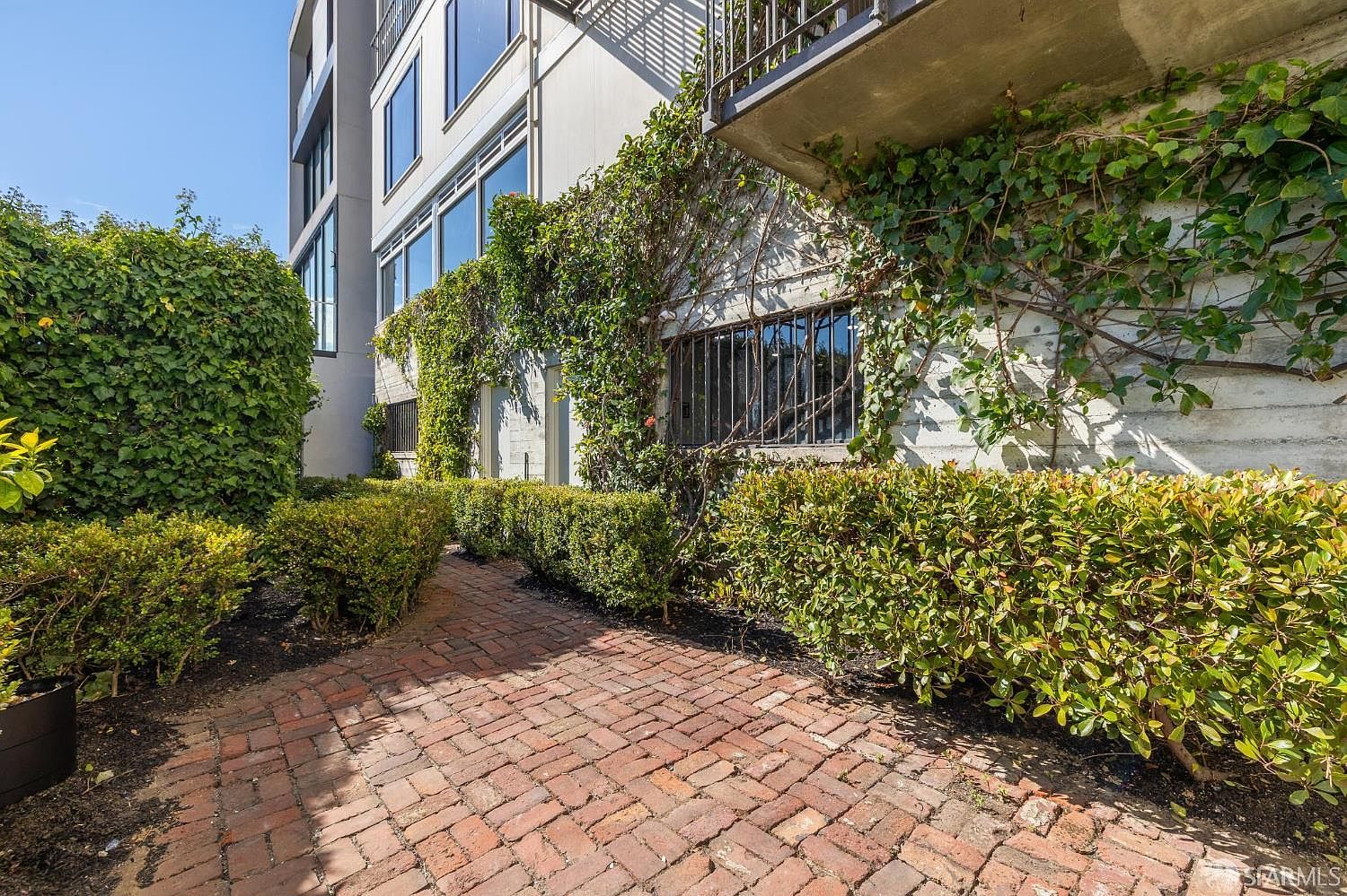 This image showcases a charming garden area with a brick pathway winding through lush greenery. The building's exterior is partially covered in climbing vines, adding a touch of natural elegance. The manicured hedges and brickwork create a well-maintained and inviting outdoor space.