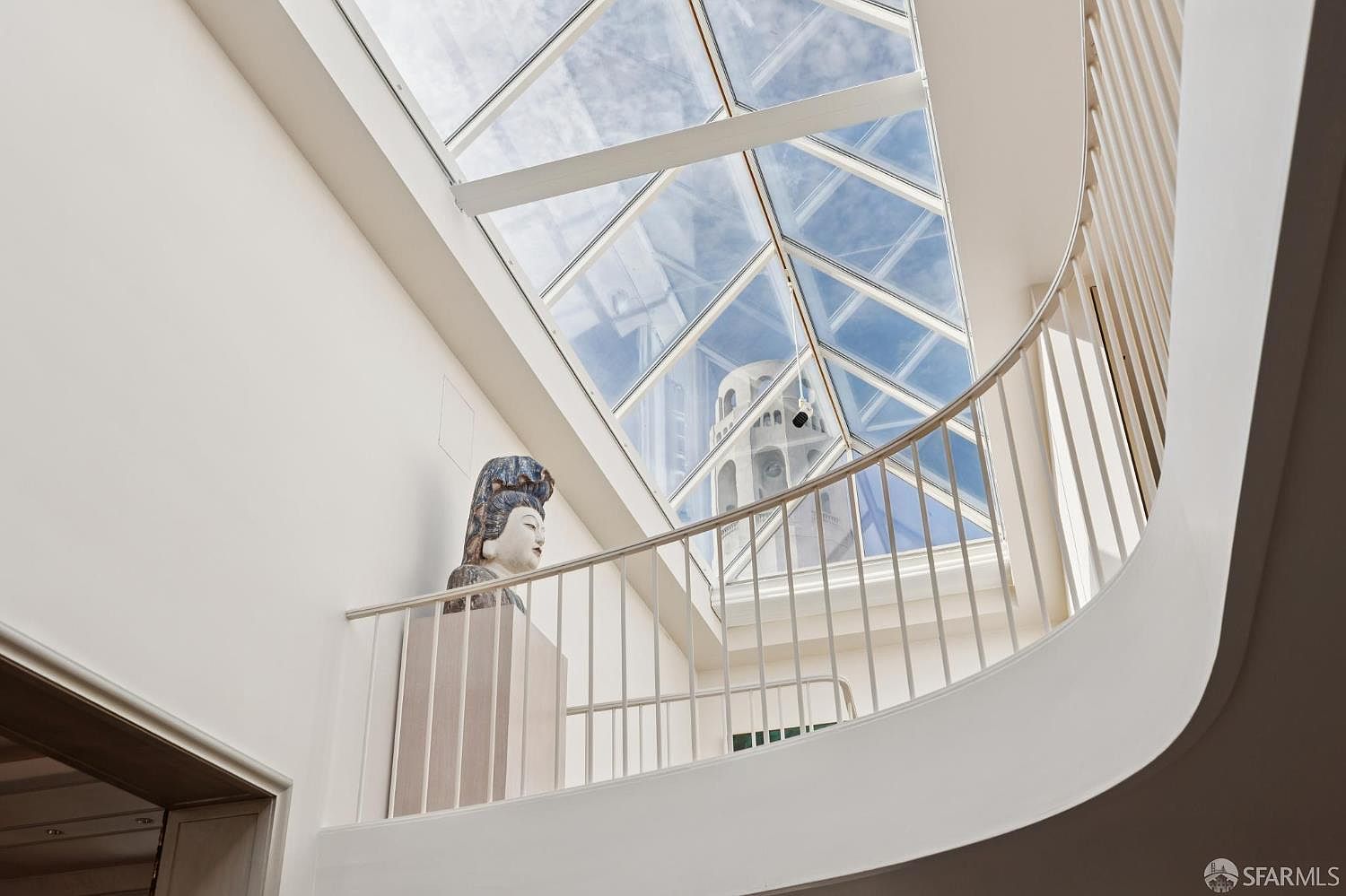 This interior shot showcases a bright, modern hallway with a unique architectural design. A curved white railing overlooks a lower level, and a striking skylight floods the space with natural light, highlighting a decorative statue. The overall impression is airy and sophisticated, emphasizing the home's distinctive features.