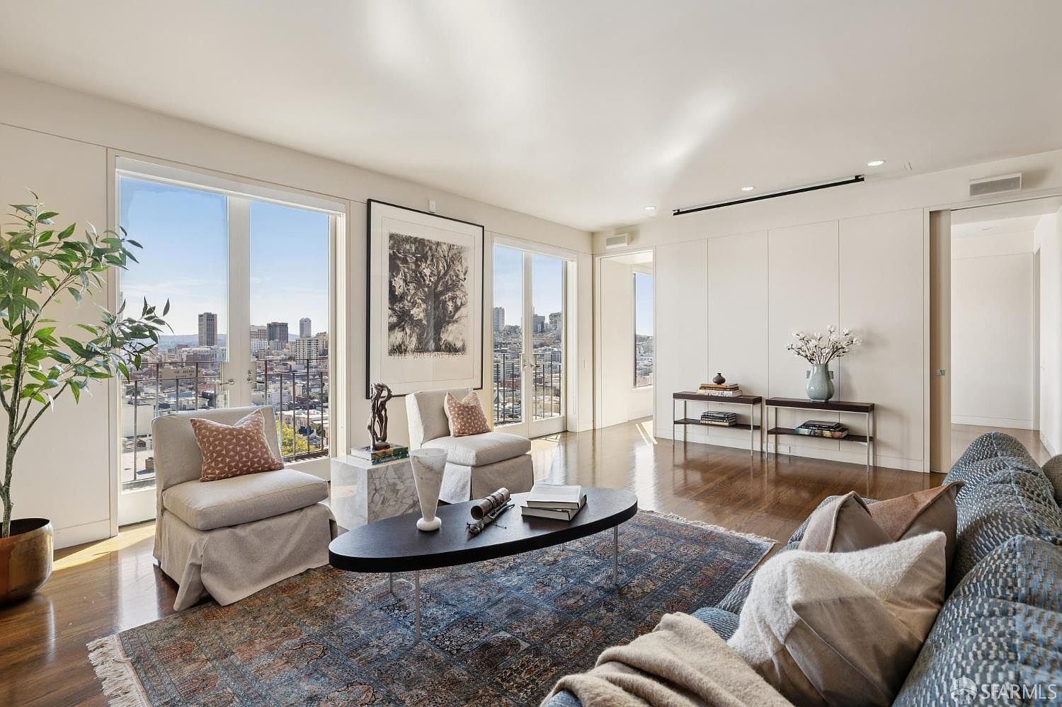 This is an interior shot of a luxurious living room featuring hardwood floors, large windows with city views, and stylish furniture. The room is decorated with a mix of modern and classic elements, including comfortable armchairs, a dark oval coffee table, and a patterned area rug. The space is bright and airy, creating an inviting and sophisticated atmosphere.
