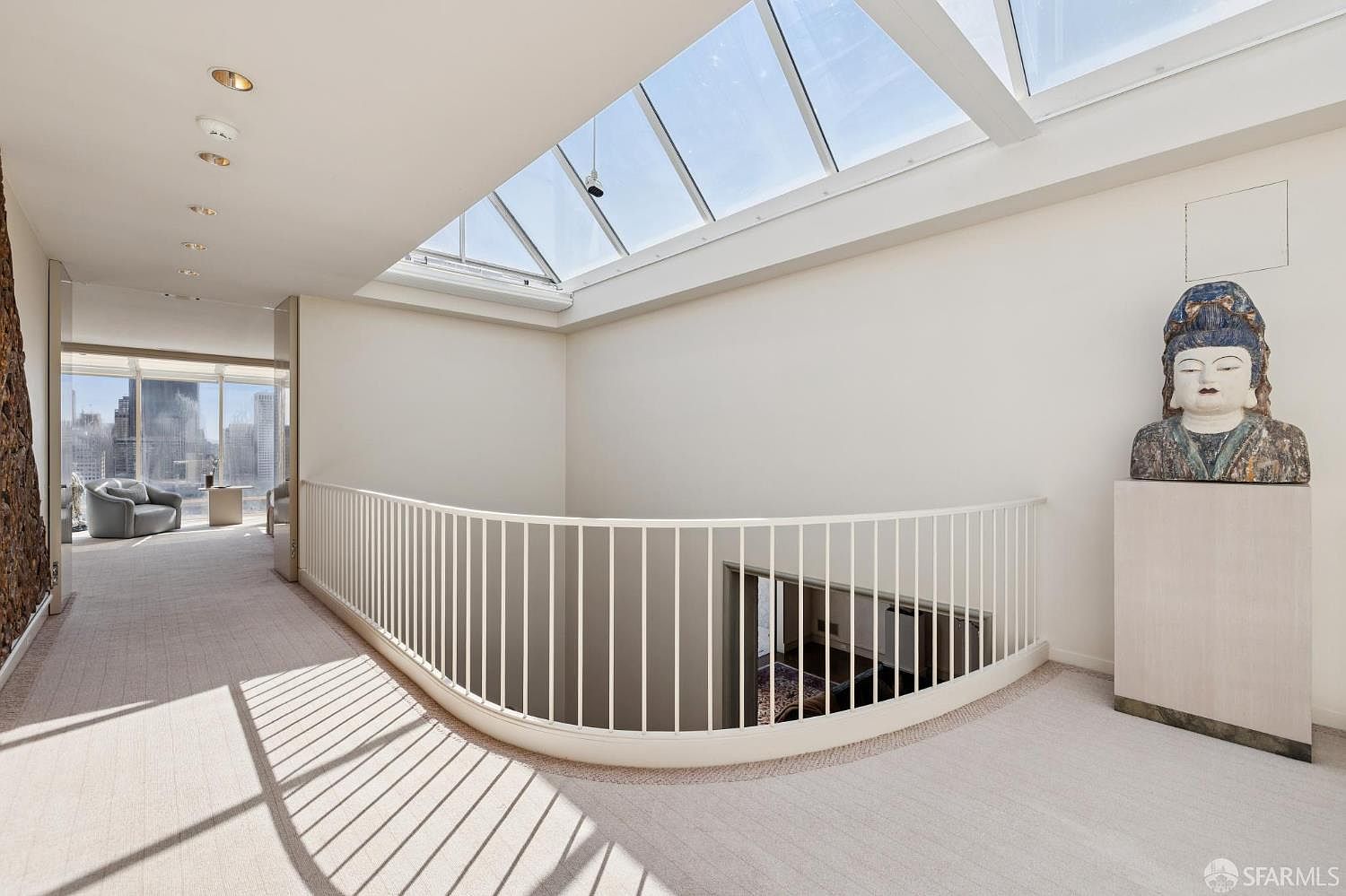 This interior shot showcases a bright hallway with a unique architectural design. A curved white railing overlooks a lower level, while a large skylight floods the space with natural light. A statue sits atop a pedestal, adding an artistic touch to the area, and a glimpse of a living area with city views can be seen in the background.