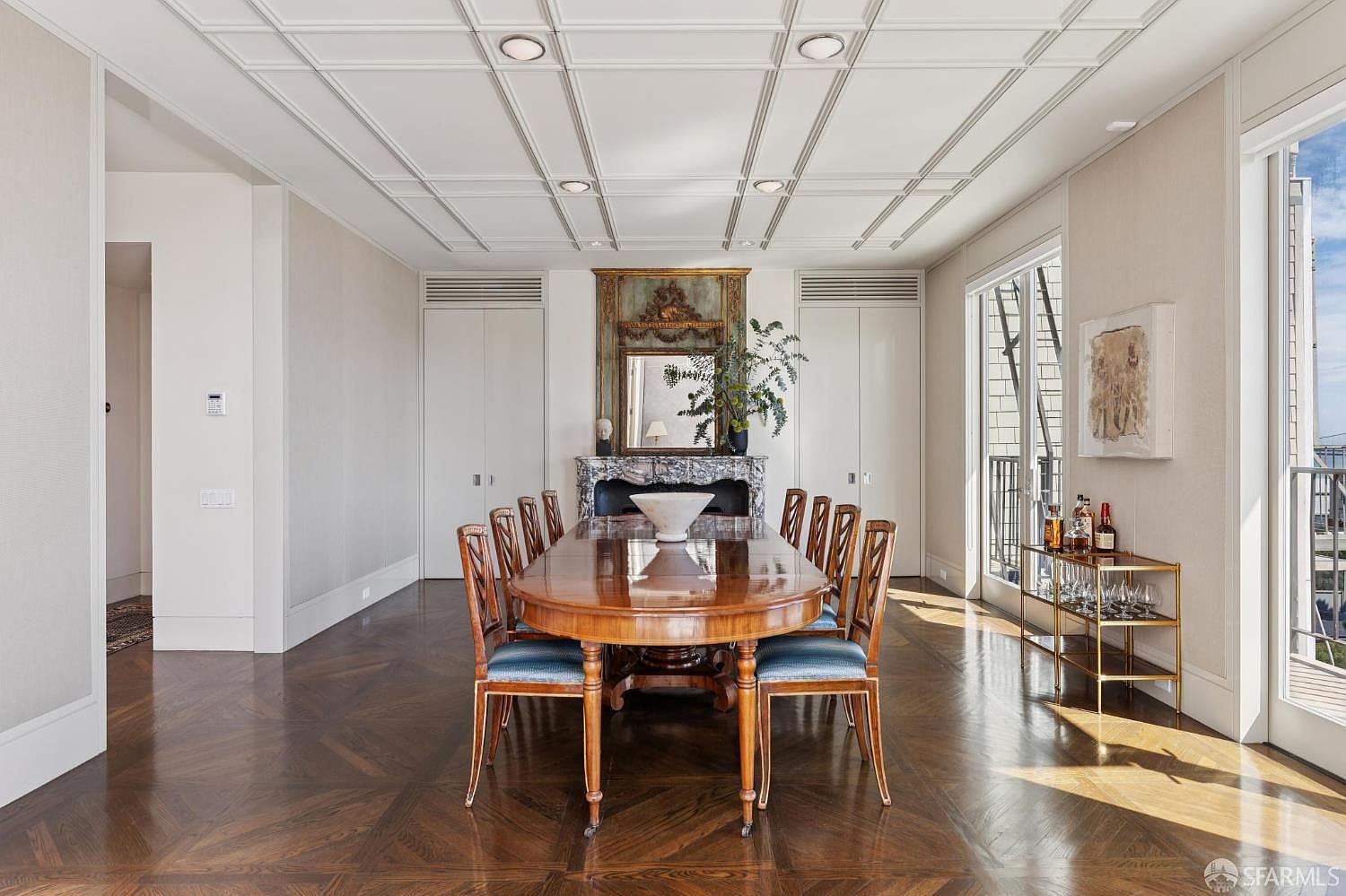 This is an interior shot of a formal dining room. The room features a large, polished wooden dining table surrounded by chairs with blue cushions, set on a herringbone-patterned hardwood floor. A decorative fireplace with a mirror above it is centered on one wall, flanked by built-in cabinets, and a bar cart is positioned near a window, suggesting an elegant and sophisticated space.