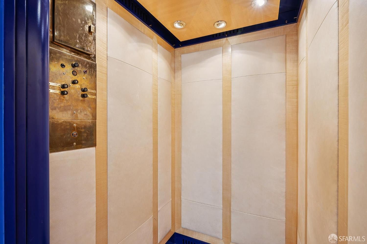 The image showcases the interior of an elevator, featuring a control panel with brass buttons and a light-colored wood-paneled interior. The design is clean and modern, with blue accents on the edges and ceiling, creating a sophisticated and functional space. The lighting is bright and even, highlighting the textures and materials used.
