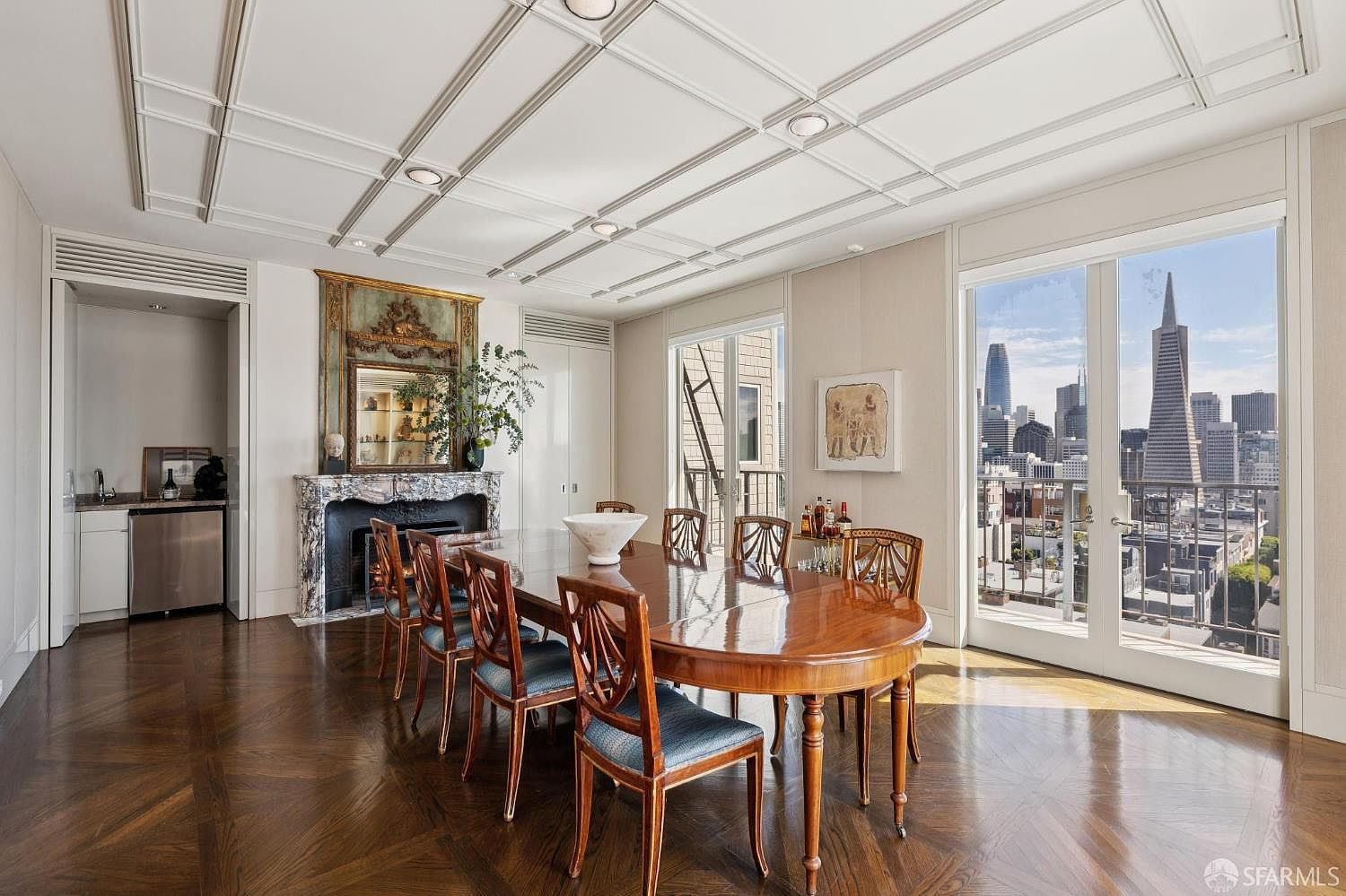 This is a dining room featuring a large, oval wooden table surrounded by elegant chairs with blue cushions. A decorative fireplace with a mirror above it adds a touch of classic charm, while large windows offer a stunning city view. The room is well-lit and features a coffered ceiling, creating a sophisticated and inviting atmosphere.