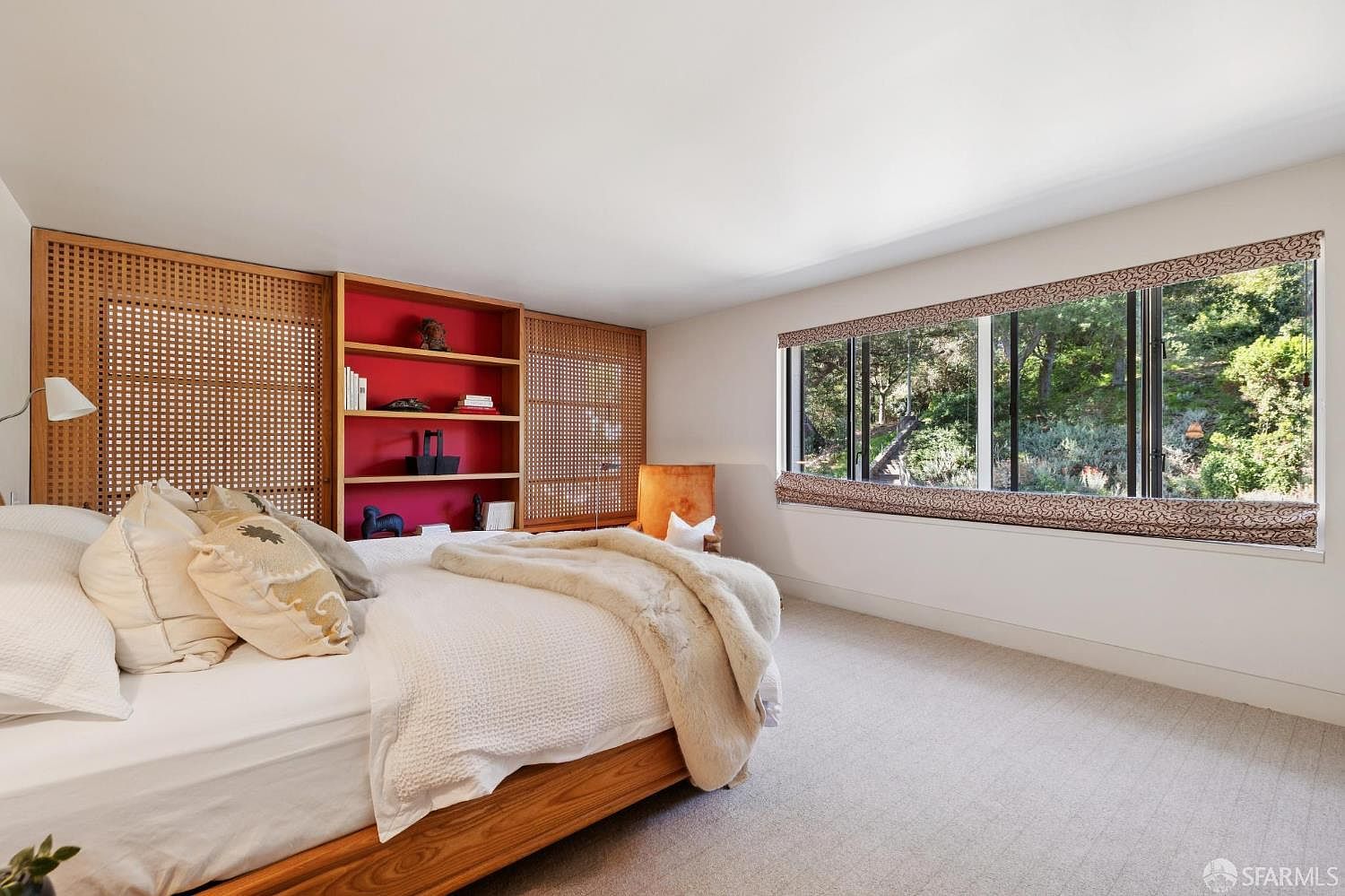 This is a bright and airy primary bedroom featuring a large window with a view of lush greenery. The room is decorated with a built-in bookshelf and a light-colored carpet. The bed is neatly made with several pillows and a soft throw blanket, creating a cozy and inviting atmosphere.