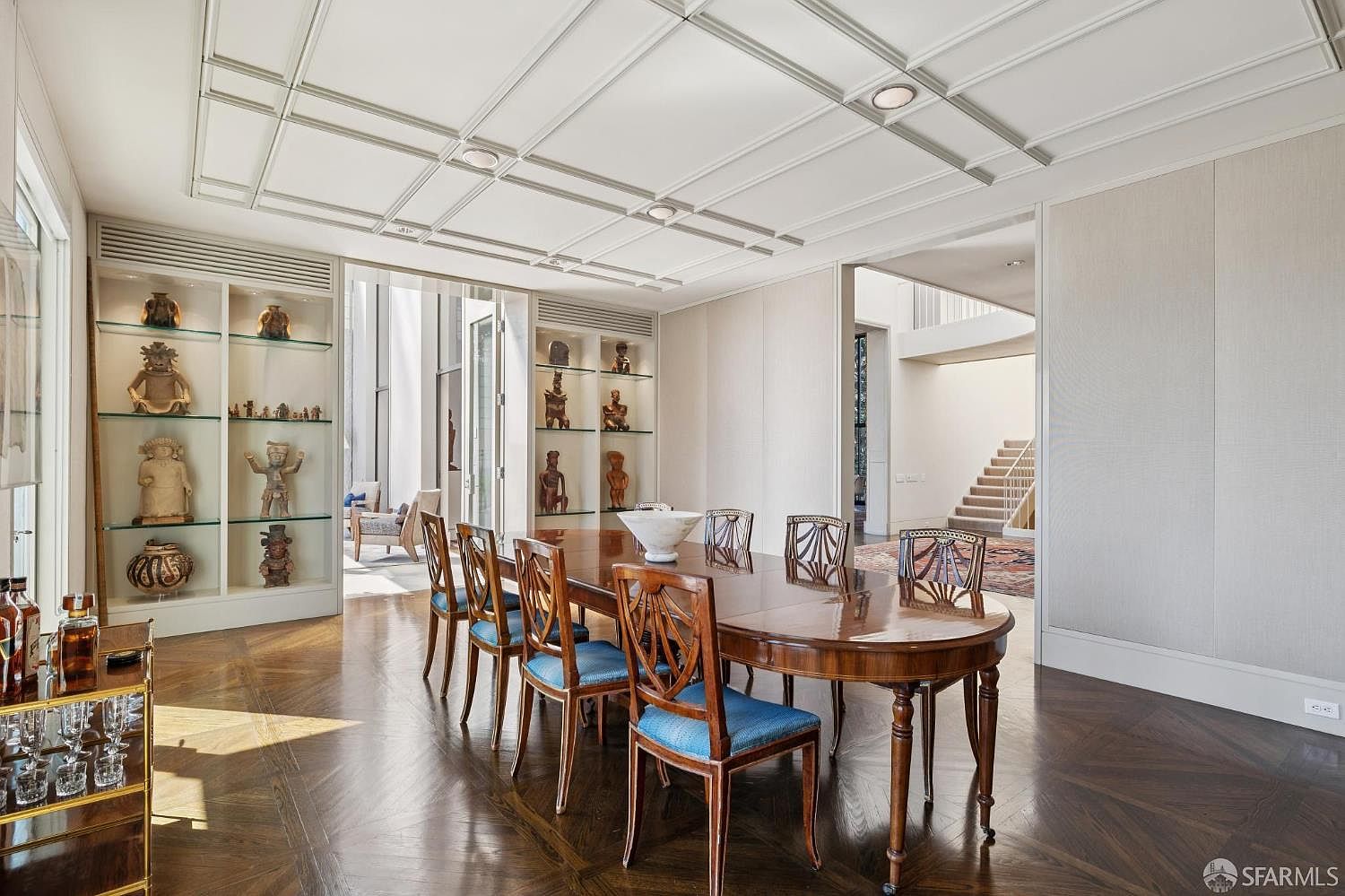 This is an interior shot of a dining room featuring a large, oval wooden dining table with blue upholstered chairs. The room has built-in display cabinets filled with artifacts, a coffered ceiling with recessed lighting, and hardwood floors in a herringbone pattern. The space is well-lit and appears elegant and spacious, with an open doorway leading to another area of the house.