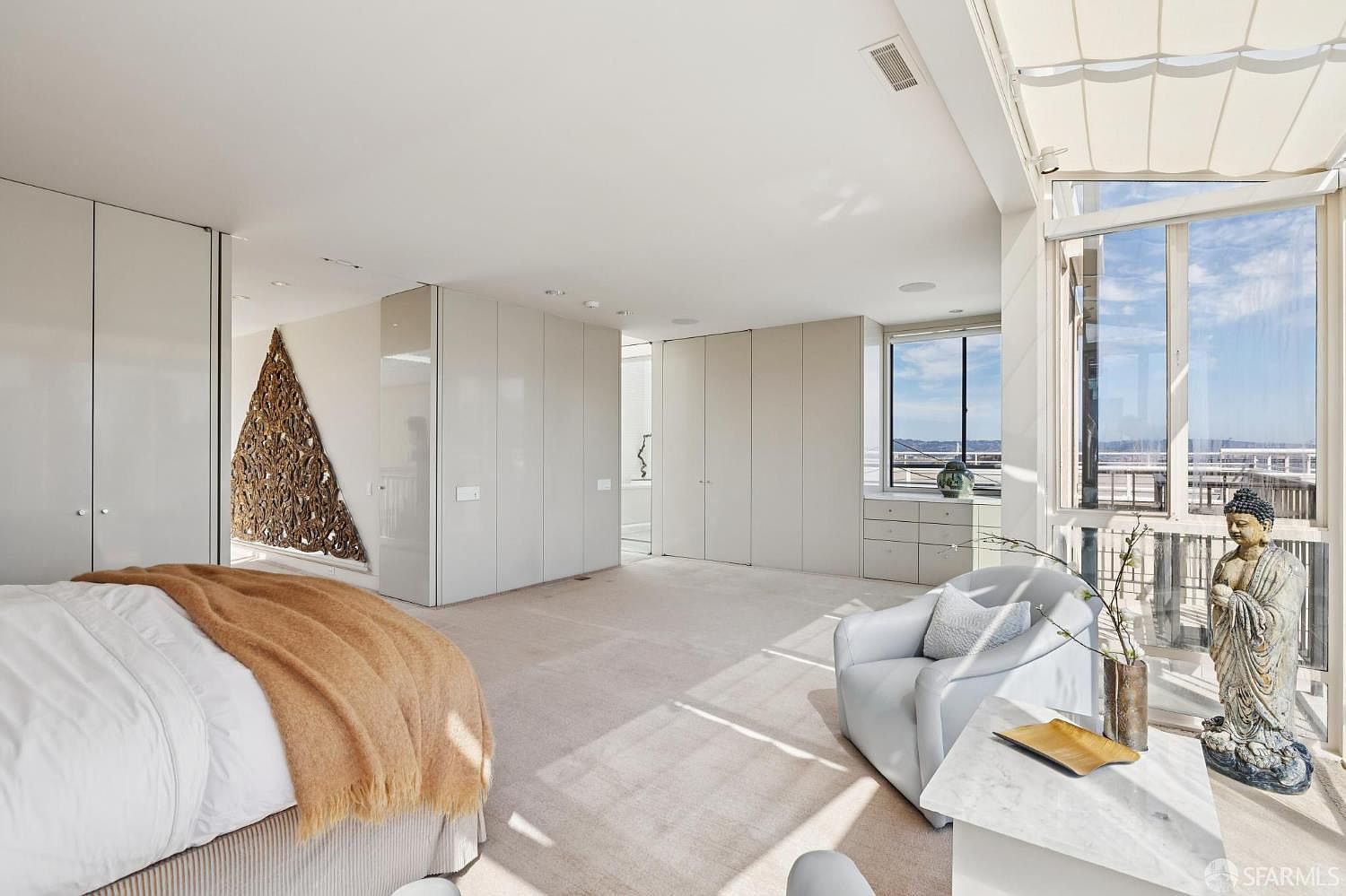 This is a spacious primary bedroom featuring a large bed with a tan throw, built-in white wardrobes, and a sitting area with a modern white armchair. The room has neutral carpet and a large window offering a view, with a Buddha statue adding a touch of serenity. The overall impression is clean, bright, and luxurious.