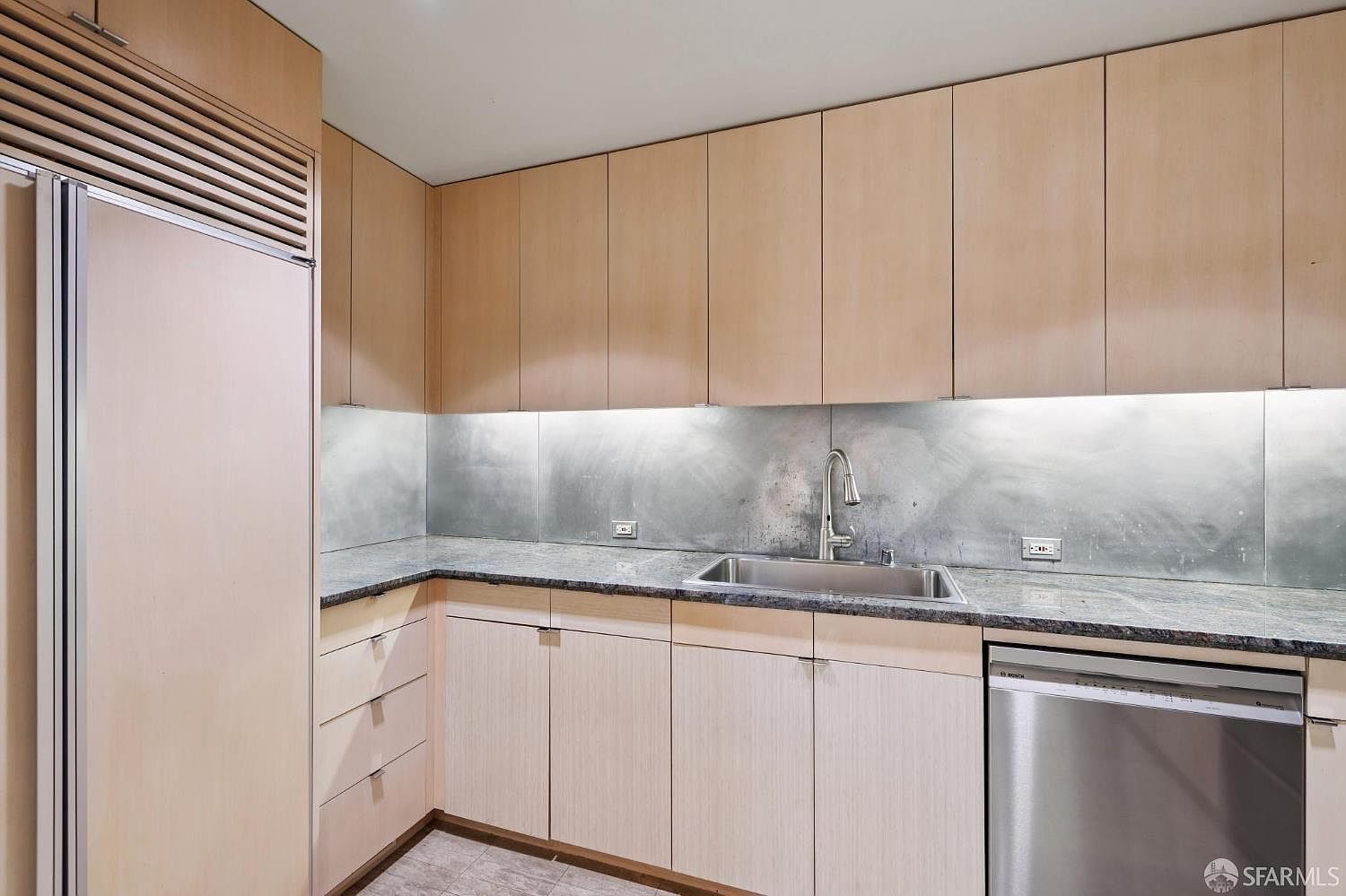 This is a well-lit kitchen featuring light wood cabinetry and stainless steel appliances. The countertops are a dark granite, and the backsplash is a metallic material. The kitchen appears modern and functional, with ample storage space.