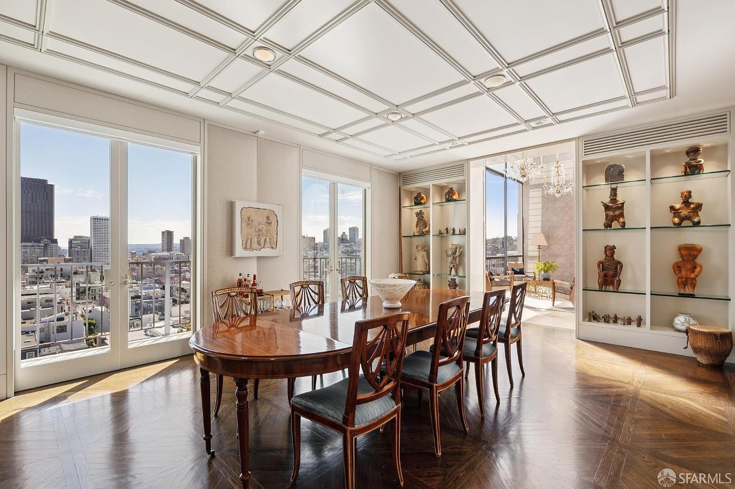 This is an elegant dining room featuring a large, oval wooden table surrounded by chairs with patterned upholstery. The room boasts a coffered ceiling, hardwood floors in a herringbone pattern, and built-in display cabinets filled with decorative objects. Large windows offer a stunning city view, enhancing the room's luxurious and sophisticated ambiance.