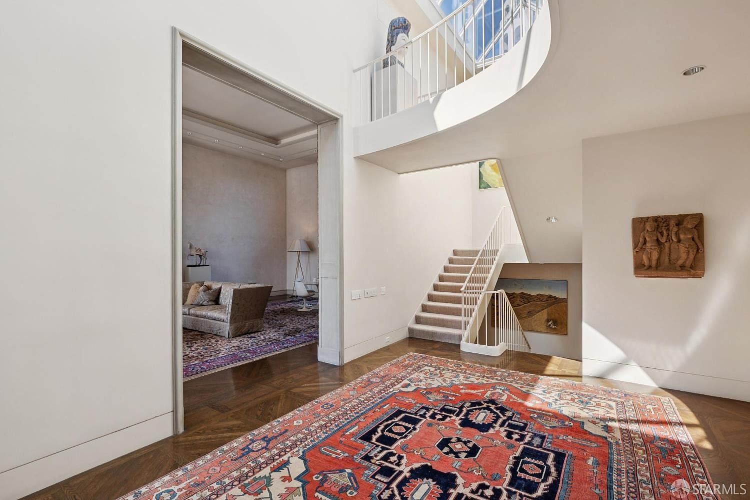 This interior shot showcases a grand hallway with a staircase. The space features hardwood floors partially covered by a large, ornate rug, white walls, and a view into a living room through a wide doorway. The staircase is carpeted and has white railings, leading to an upper level with a curved balcony, creating a bright and airy atmosphere.
