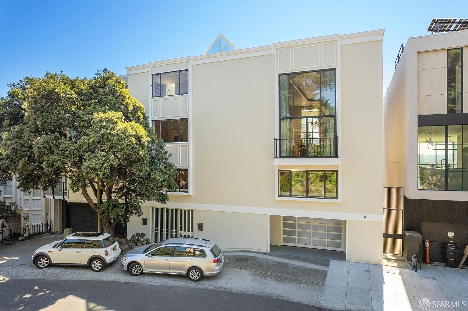 This is a front exterior view of a modern multi-story home. The facade is light-colored with large windows and a balcony. Two cars are parked in front of the house, and a mature tree adds to the curb appeal. The overall impression is of a well-maintained and stylish property.