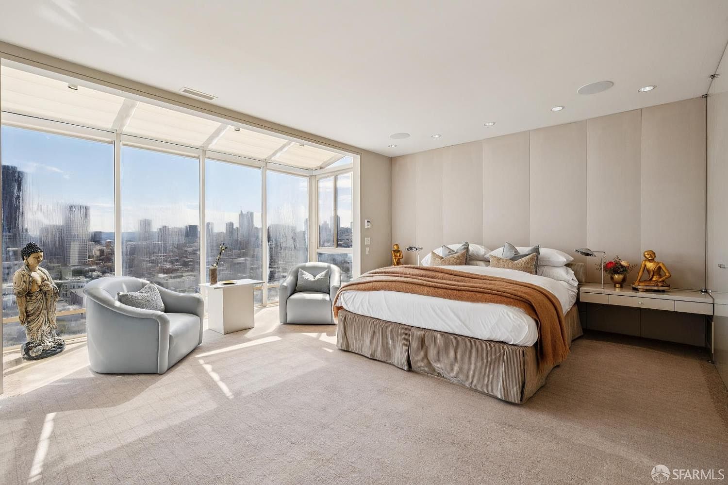 This is a luxurious primary bedroom featuring floor-to-ceiling windows with a stunning city view. The room is decorated in neutral tones with a large bed, two armchairs, and decorative statues. The overall impression is one of elegance and tranquility, perfect for a high-end real estate listing.