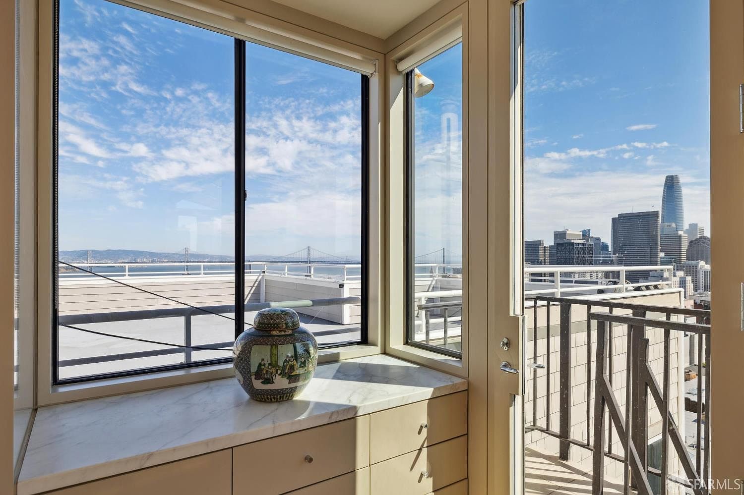 This sunroom features large windows offering expansive views of the city skyline and the bay with the bridge. The room includes built-in cabinetry with a marble countertop, adding a touch of elegance. The space is bright and airy, creating a serene atmosphere.