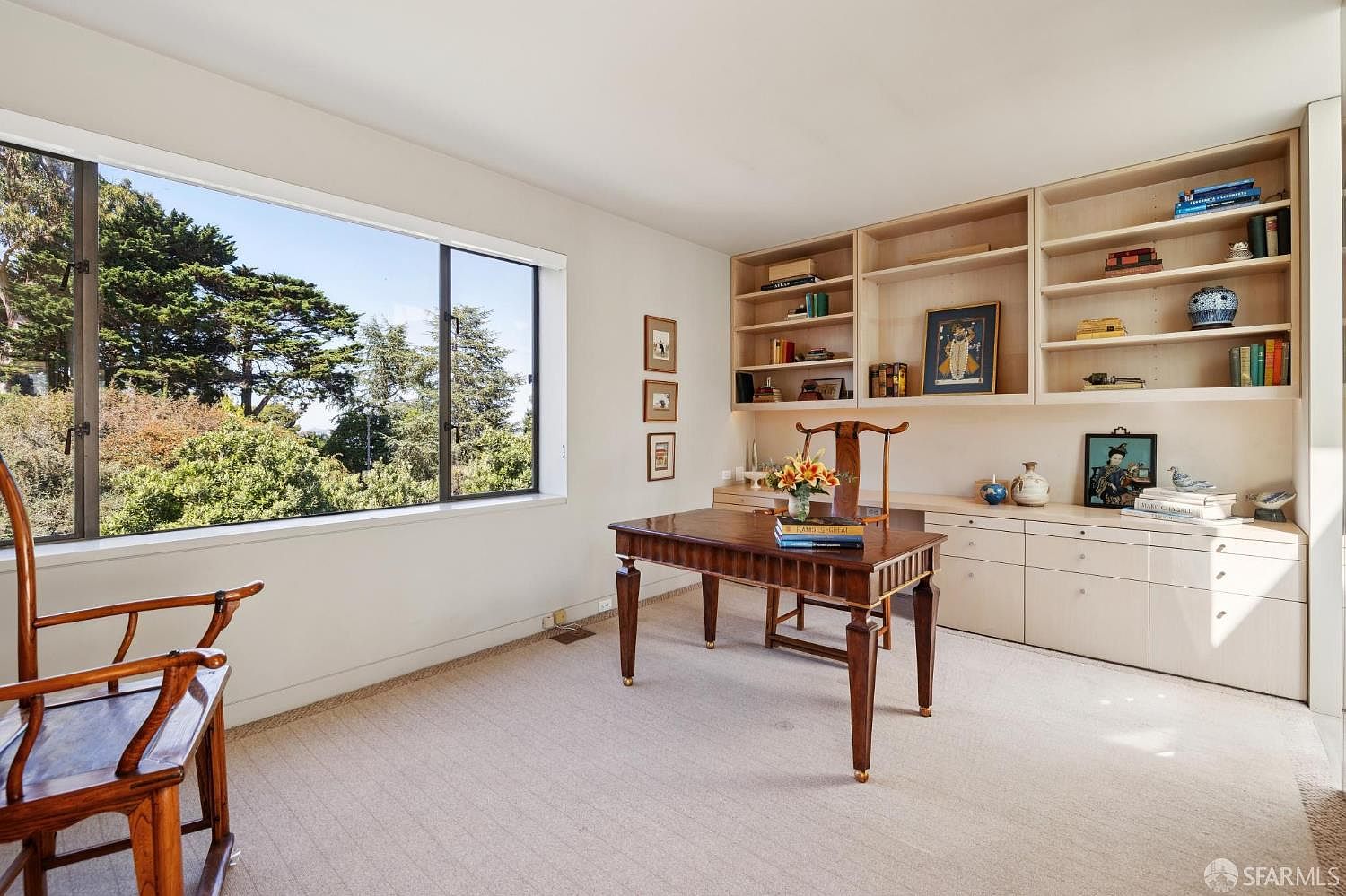 This is an interior shot of a well-lit office or study. The room features a large window with a view of lush greenery, built-in shelving and cabinets, and a wooden desk with a chair. The overall impression is one of a calm and organized workspace.