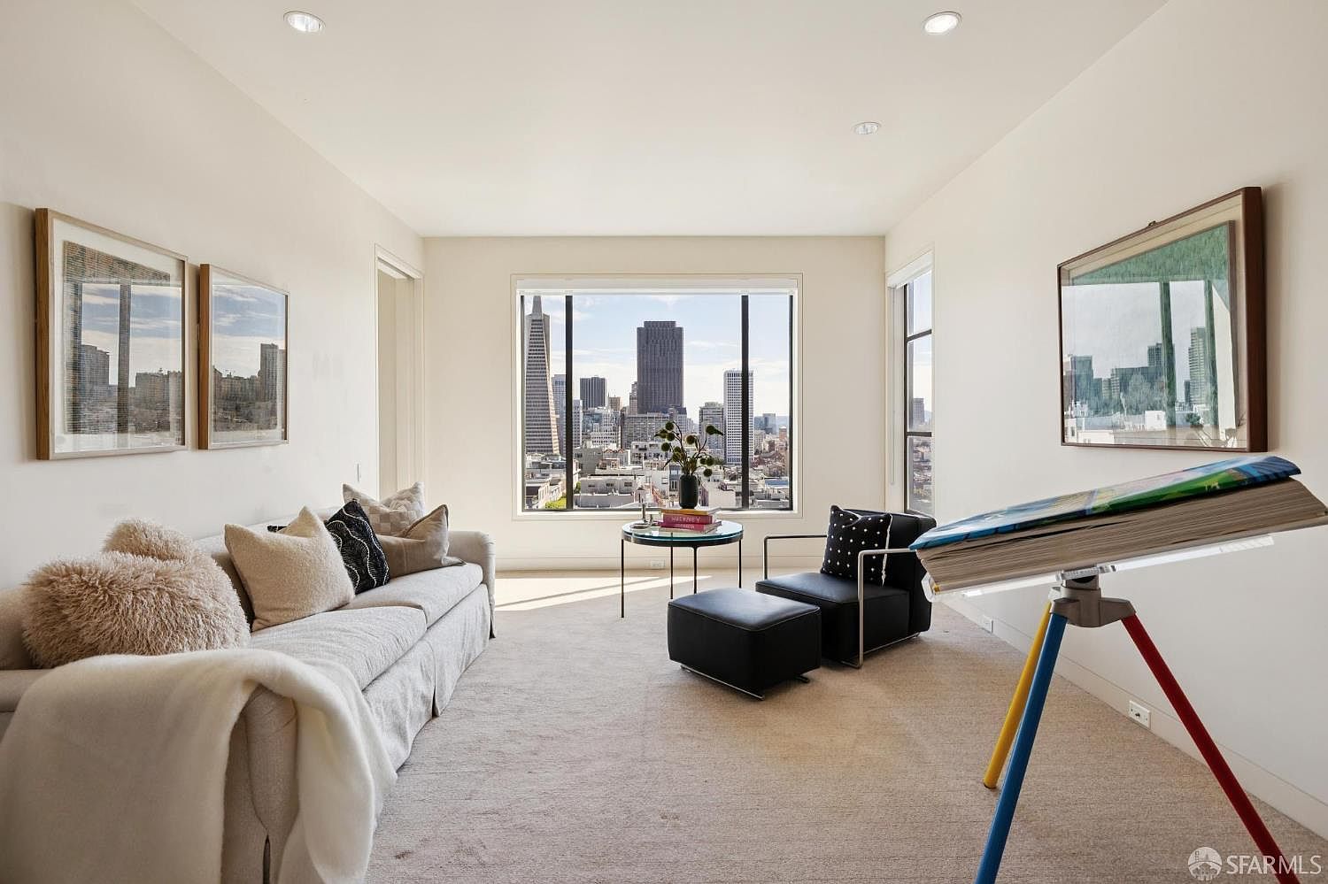 This is an interior shot of a living room featuring a neutral color palette with a comfortable sofa adorned with various pillows and a throw blanket. A large window provides a stunning city view, while modern furniture and artwork add a touch of sophistication. The room is well-lit and appears spacious, creating an inviting atmosphere.