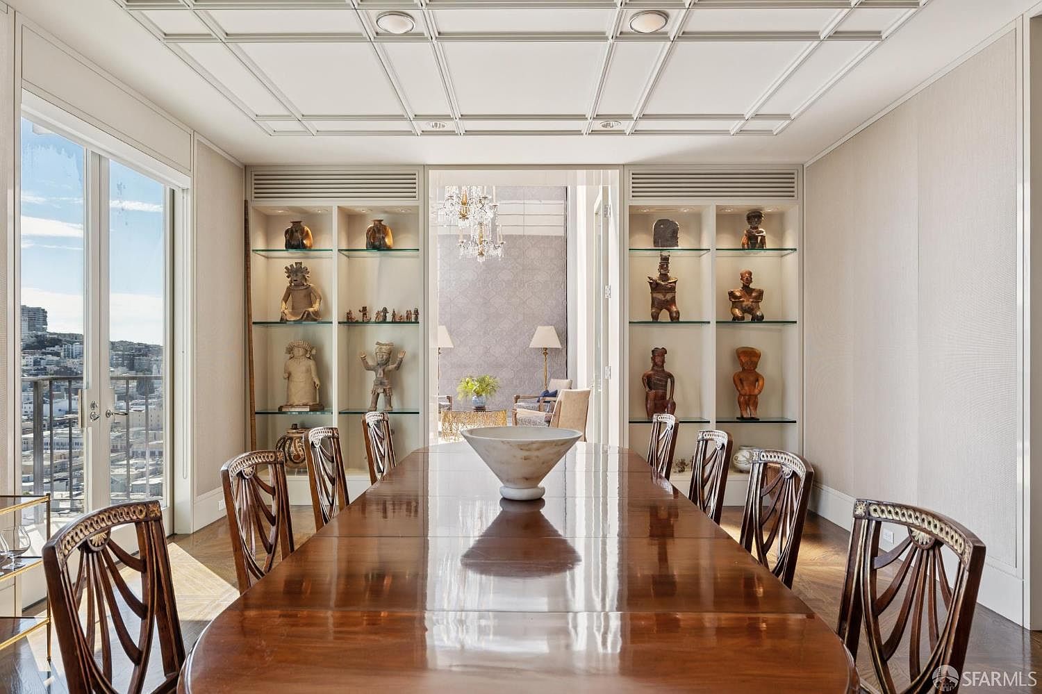 This is an interior shot of a dining room featuring a long, polished wooden table surrounded by ornate chairs. Built-in shelving units display decorative objects, and a large window offers a city view. The room is well-lit and exudes a sense of elegance and sophistication.