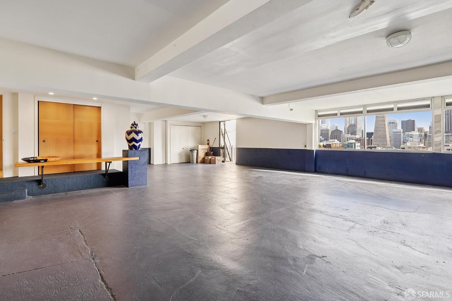 This is a spacious, modern living room with a large window offering a city view. The room features a polished concrete floor, white walls, and exposed beams, creating an open and airy atmosphere. A built-in bench and a decorative vase add unique architectural details to the space.