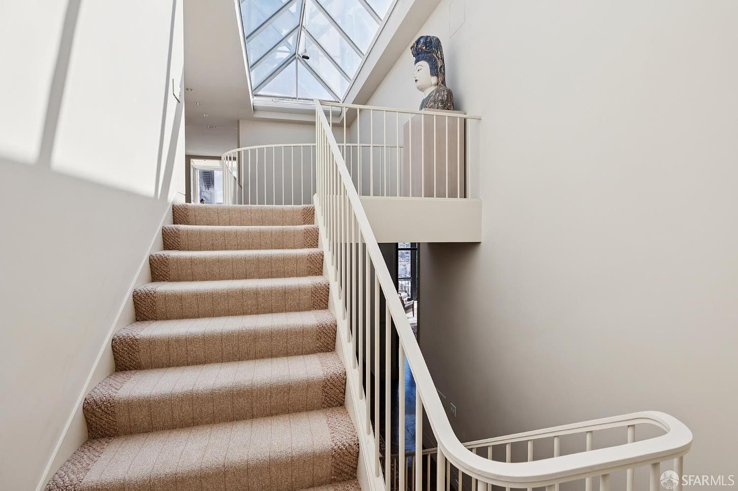 This interior shot showcases a carpeted staircase leading upwards, bathed in natural light from a skylight above. A white railing curves along the stairs and continues onto a small landing where a decorative statue is placed. The neutral color palette and architectural details create a bright and inviting atmosphere.