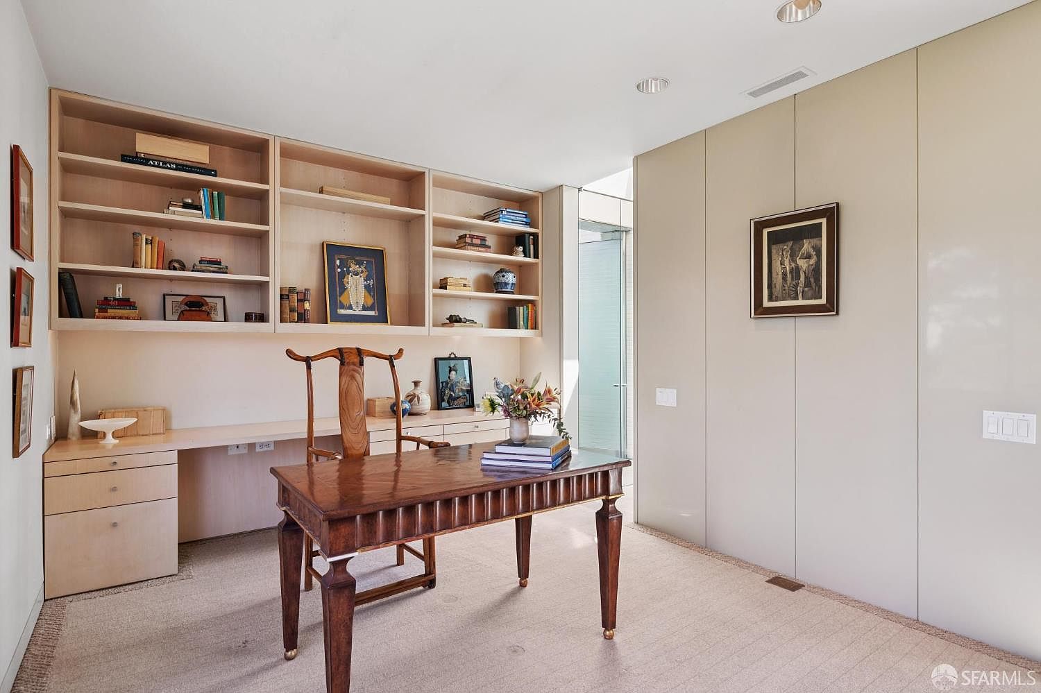 This is an interior shot of a home office or study. The room features built-in shelving with books and decorative items, a desk area, and a separate wooden desk with a chair. The walls are a neutral color, and there is artwork on the walls, creating a sophisticated and functional workspace.