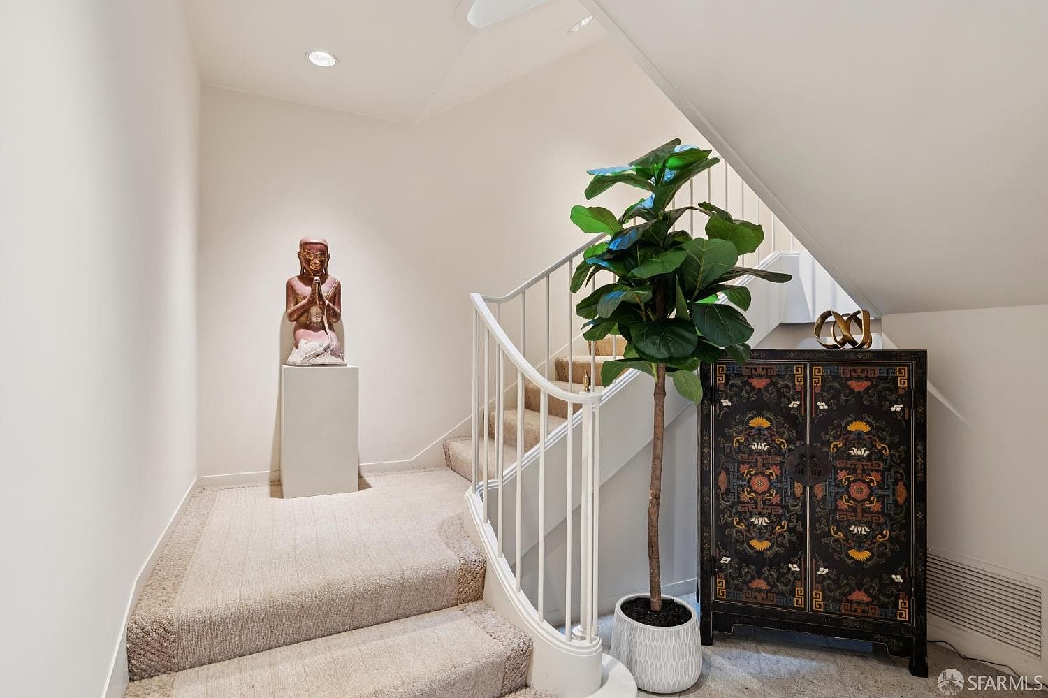 This interior shot showcases a well-lit hallway with carpeted stairs. A statue sits atop a pedestal on the left, while a large potted fiddle-leaf fig tree and a decorative cabinet adorn the right side of the staircase. The white walls and white stair railing create a clean and modern aesthetic.