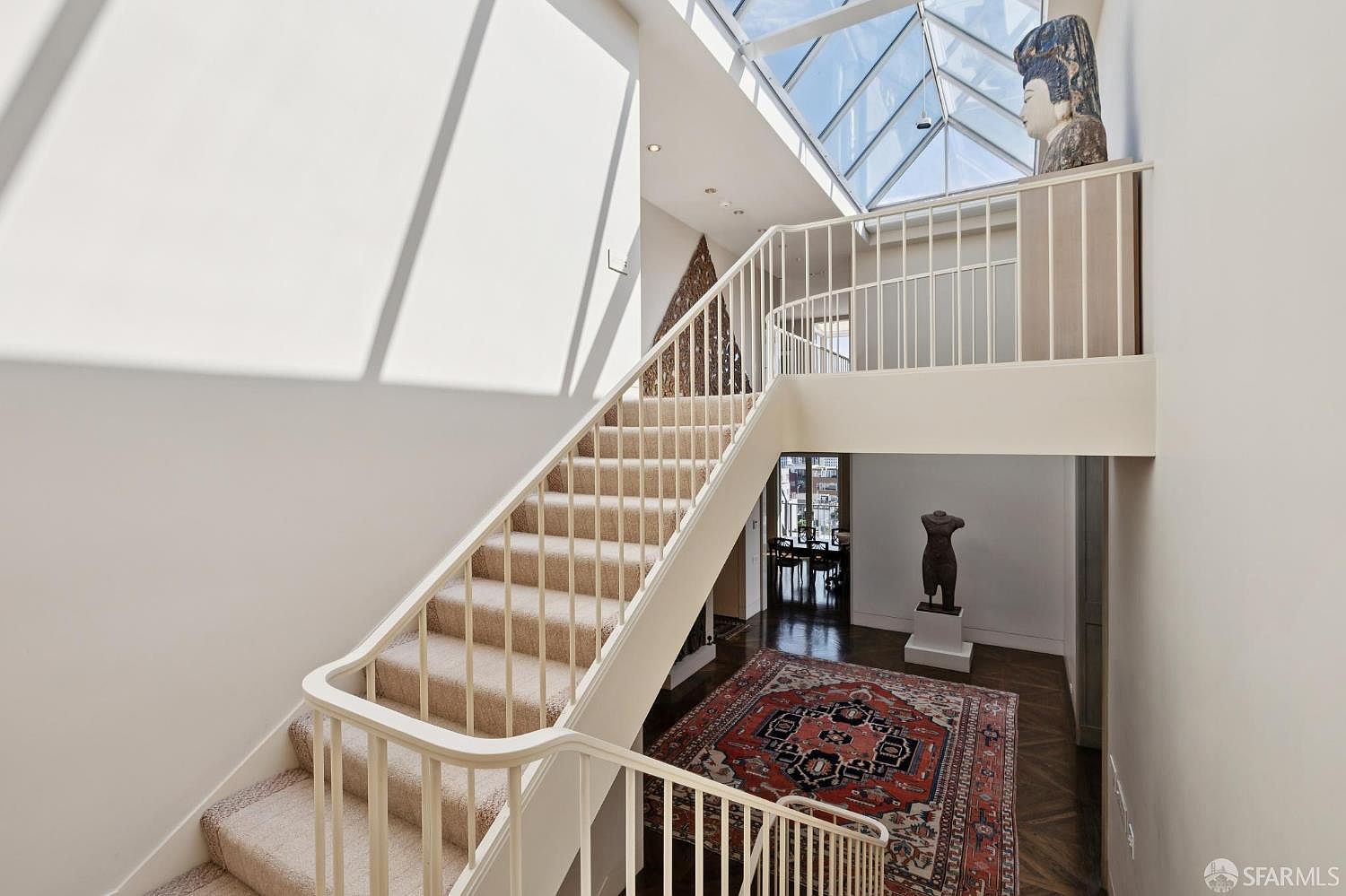 This interior shot showcases a grand hallway and staircase bathed in natural light from a skylight. The staircase is carpeted with a light-colored material and features white railings. A decorative rug adorns the hardwood floor at the base of the stairs, and sculptures add an artistic touch to the space, creating an elegant and inviting atmosphere.