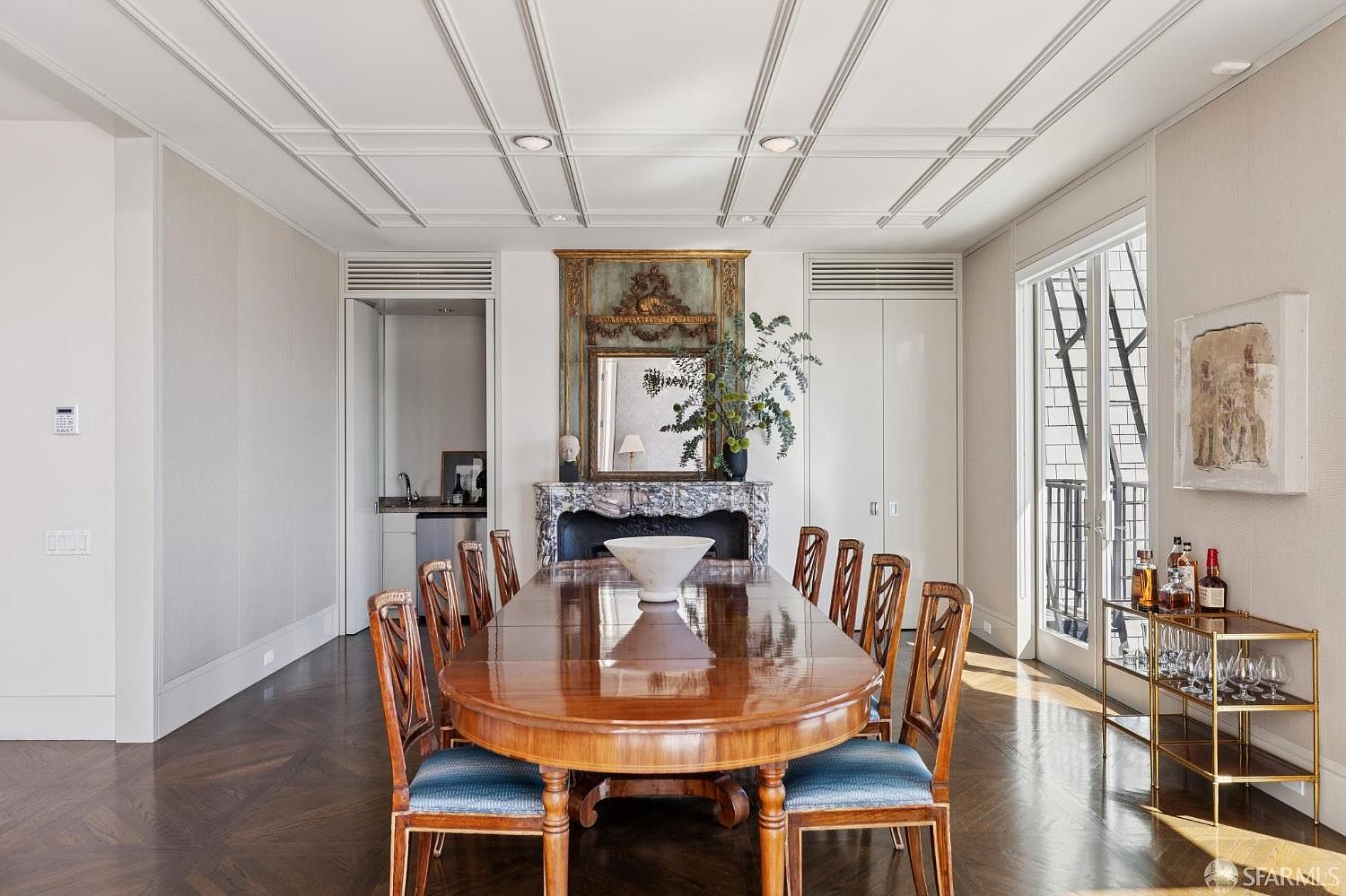 This is a dining room featuring a large, oval wooden table surrounded by chairs with blue cushions. A decorative fireplace with a mirror above it serves as a focal point, and a bar cart with bottles and glassware is visible to the right. The room has a sophisticated and elegant feel, enhanced by the herringbone wood flooring and coffered ceiling.