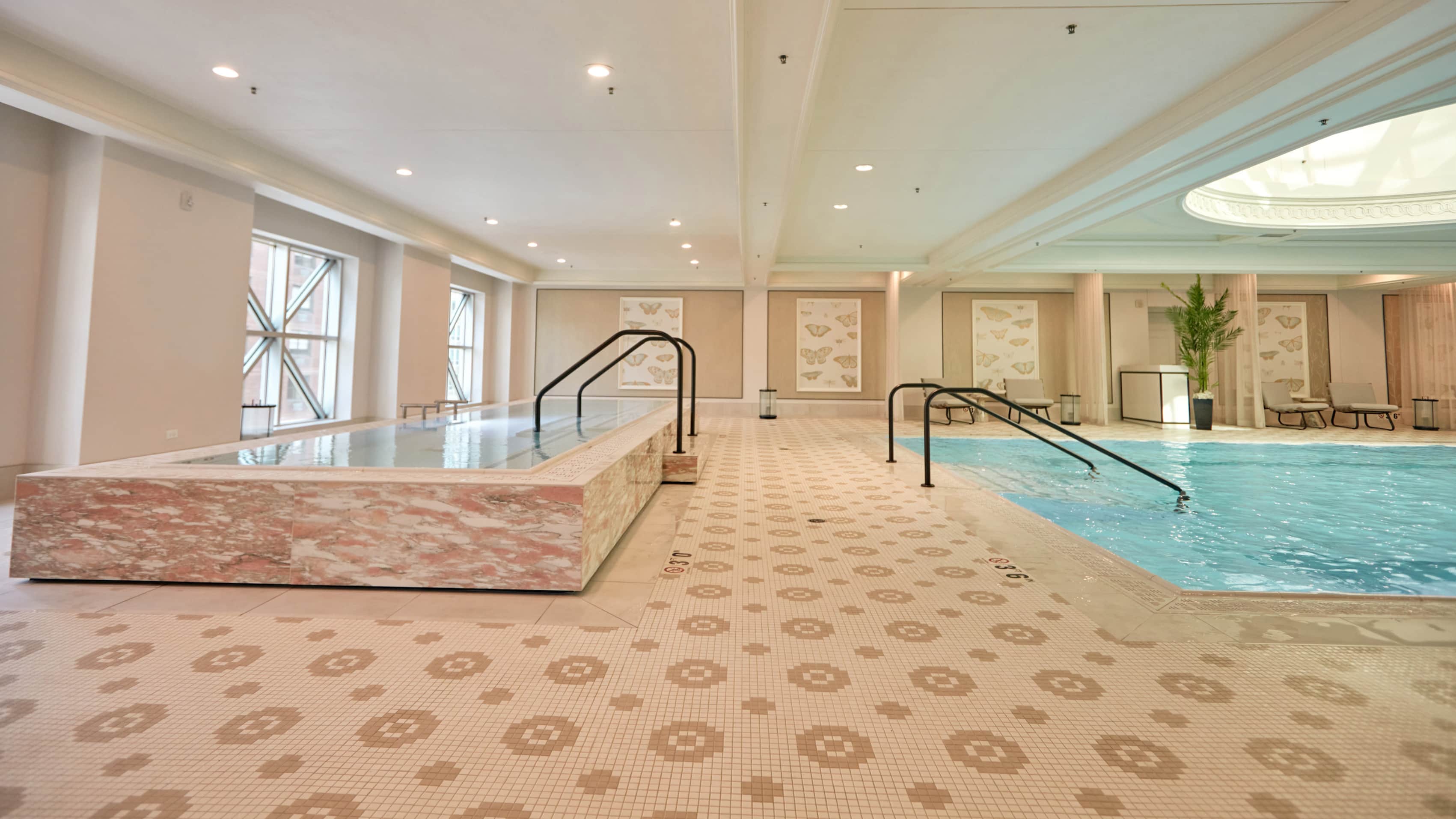 This is an interior shot of a well-lit, luxurious indoor pool and spa area. It features a hot tub with a pink marbled exterior and a larger pool with clear blue water. Decorative tile flooring and elegant artwork contribute to a high-end, relaxing atmosphere, ideal for a wellness or resort-style real estate listing.