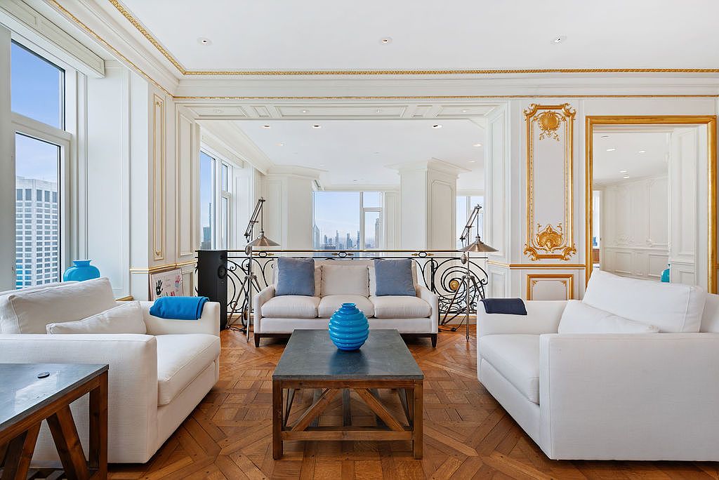 This is an elegant living room featuring white sofas, a dark wood coffee table, and parquet flooring. The room has ornate gold detailing on the walls and ceiling, with large windows offering city views. The space is bright and luxurious, creating a sophisticated atmosphere.