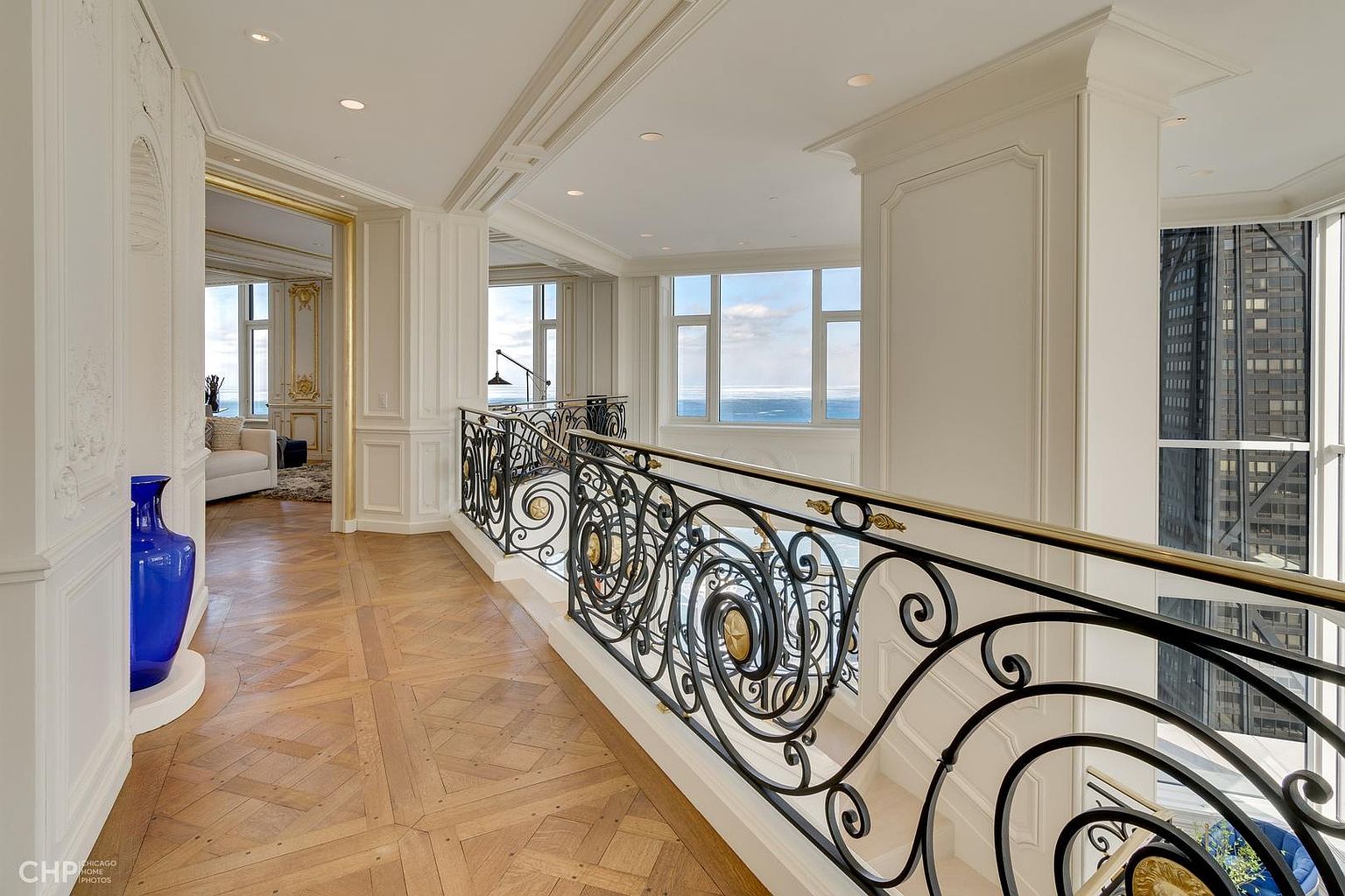 This interior shot showcases an elegant hallway with a decorative wrought iron railing and a polished parquet floor. The hallway leads to a bright living area with large windows offering a view of the water. The architectural details, including the ornate moldings and the blue vase, contribute to a luxurious and sophisticated ambiance.