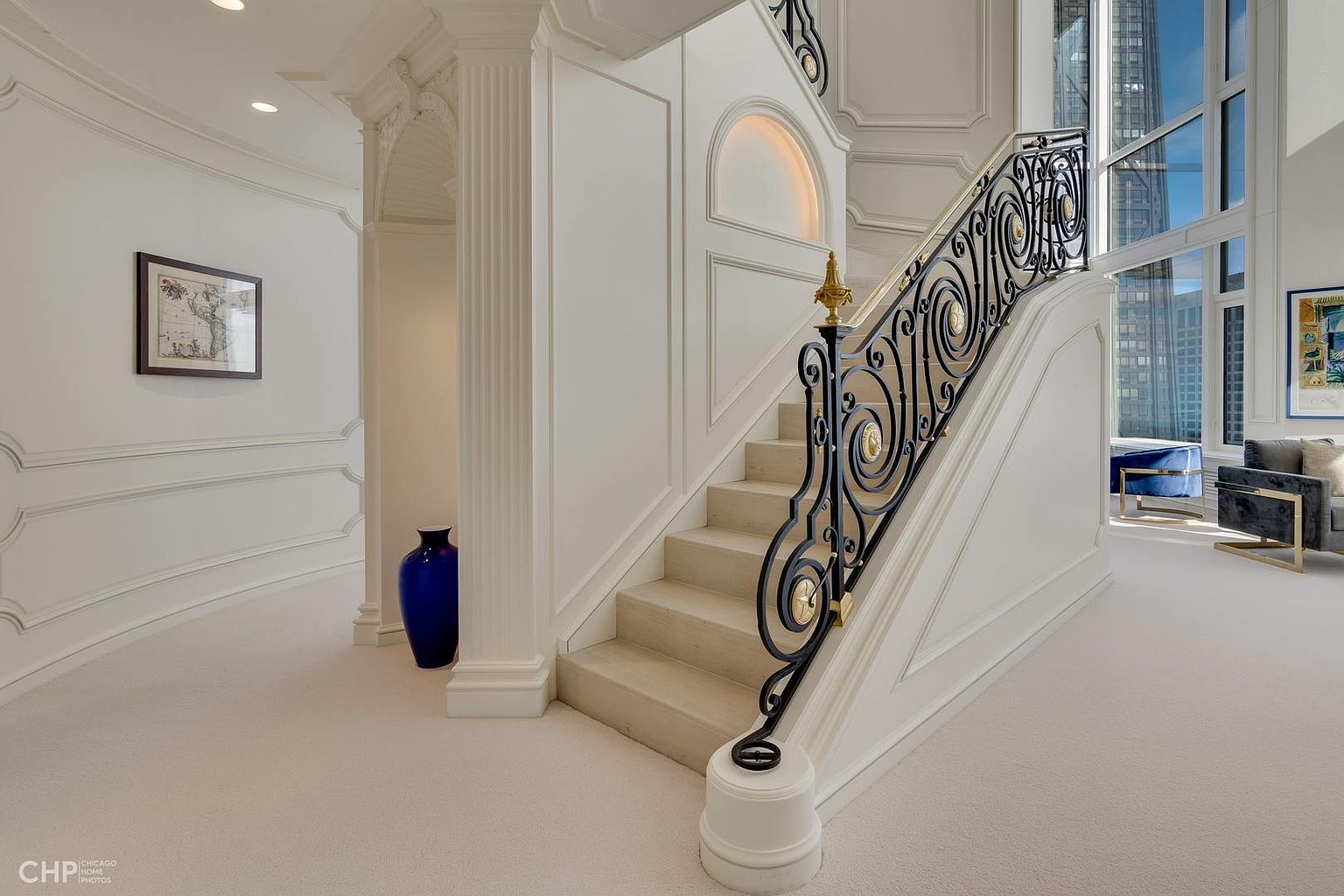 This interior shot showcases an elegant hallway and staircase. The staircase features a decorative black and gold railing, light-colored steps, and white paneled walls. The hallway includes detailed molding, a blue vase, and a framed picture, creating a luxurious and sophisticated atmosphere.