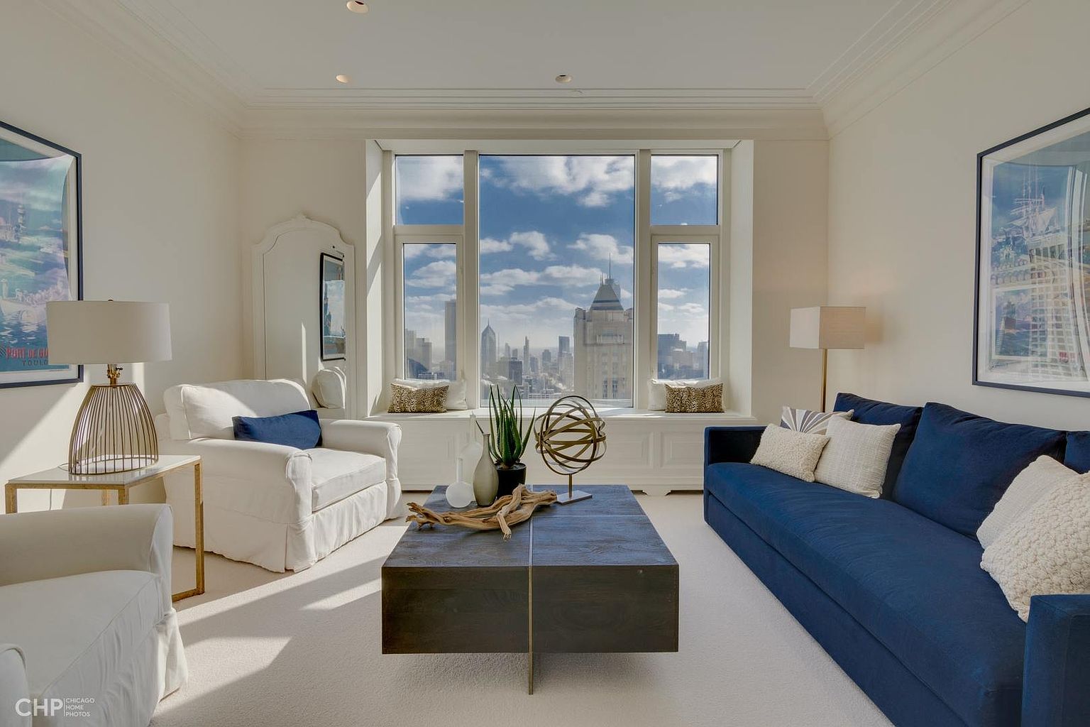 This is a bright and airy living room featuring a large window with a city view. The room is furnished with a blue sofa, white armchairs, and a dark wood coffee table. Artwork adorns the walls, and the overall style is elegant and modern, creating a welcoming and sophisticated atmosphere.