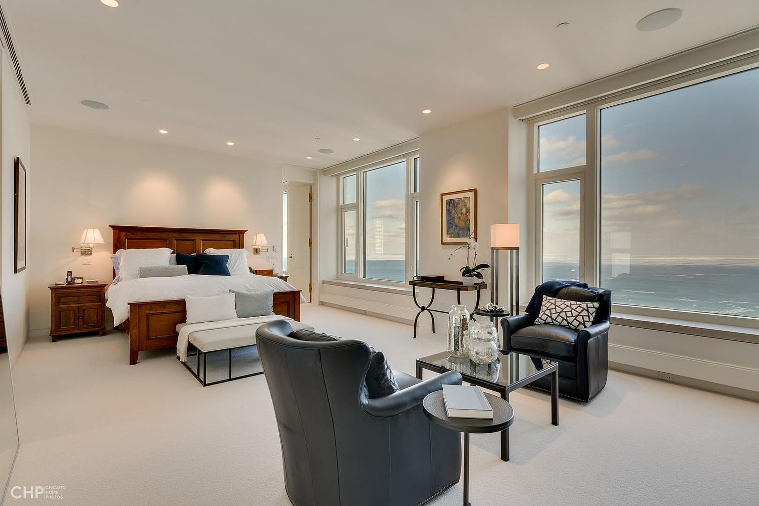 This is a luxurious primary bedroom featuring a wooden bed frame with white linens and decorative pillows. The room has large windows offering stunning ocean views, complemented by comfortable seating areas with leather armchairs and a glass coffee table. The overall impression is one of elegance and tranquility, perfect for a high-end real estate listing.