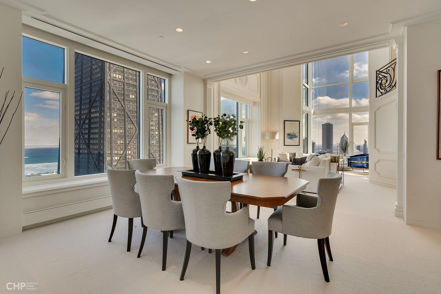 This is an interior shot of a dining room featuring a large wooden table surrounded by six upholstered chairs with dark wood legs. Three black vases with greenery sit in the center of the table. Large windows offer views of the city skyline and lake, enhancing the room's appeal. The room transitions into a living area with a view of the city skyline.