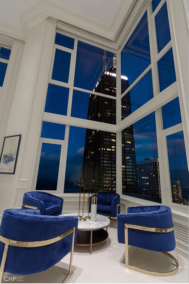 This interior shot showcases a luxurious living room with floor-to-ceiling windows offering a stunning view of a skyscraper at night. The room is furnished with stylish blue velvet chairs with gold accents and a modern round coffee table. The overall impression is one of elegance and sophistication, highlighting the property's prime location and upscale design.
