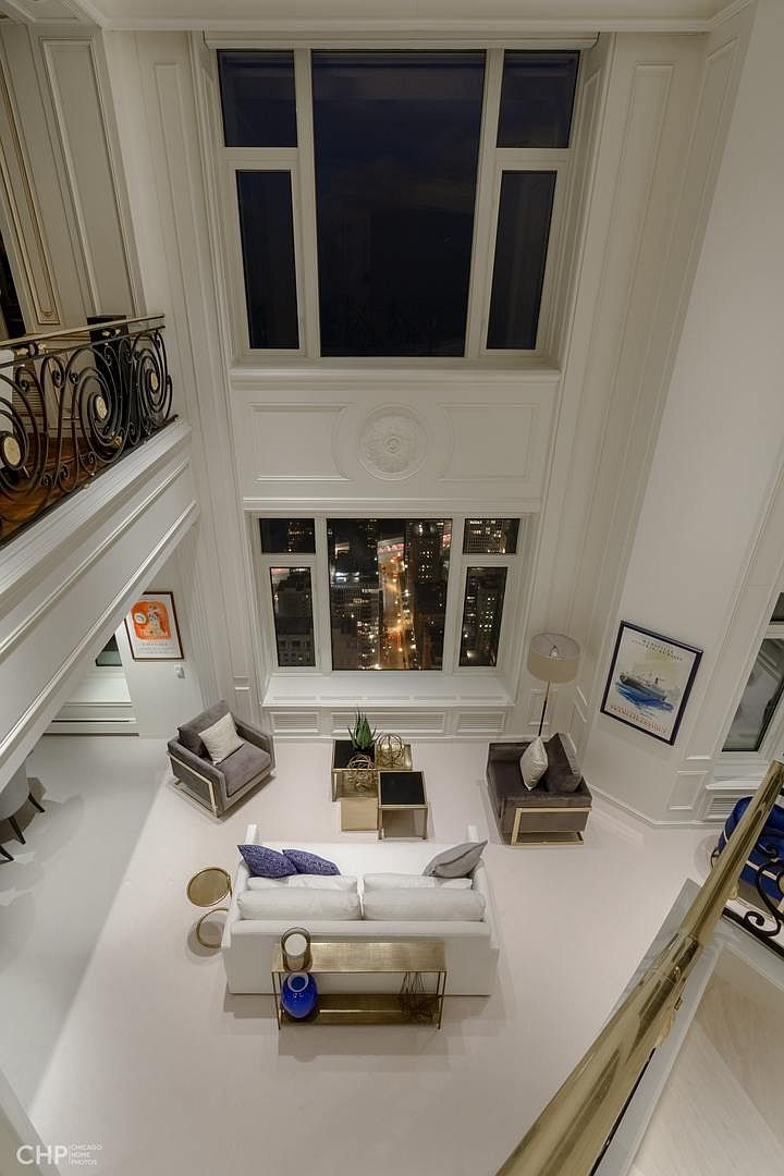 This is a high-angle shot of a luxurious living room featuring a white sofa with blue and gray pillows, a gray armchair, and a brown armchair. The room is bathed in natural light from large windows offering a city view, complemented by modern gold-accented tables and a blue vase. The overall impression is one of elegance and sophistication, highlighting the spaciousness and upscale design of the living area.